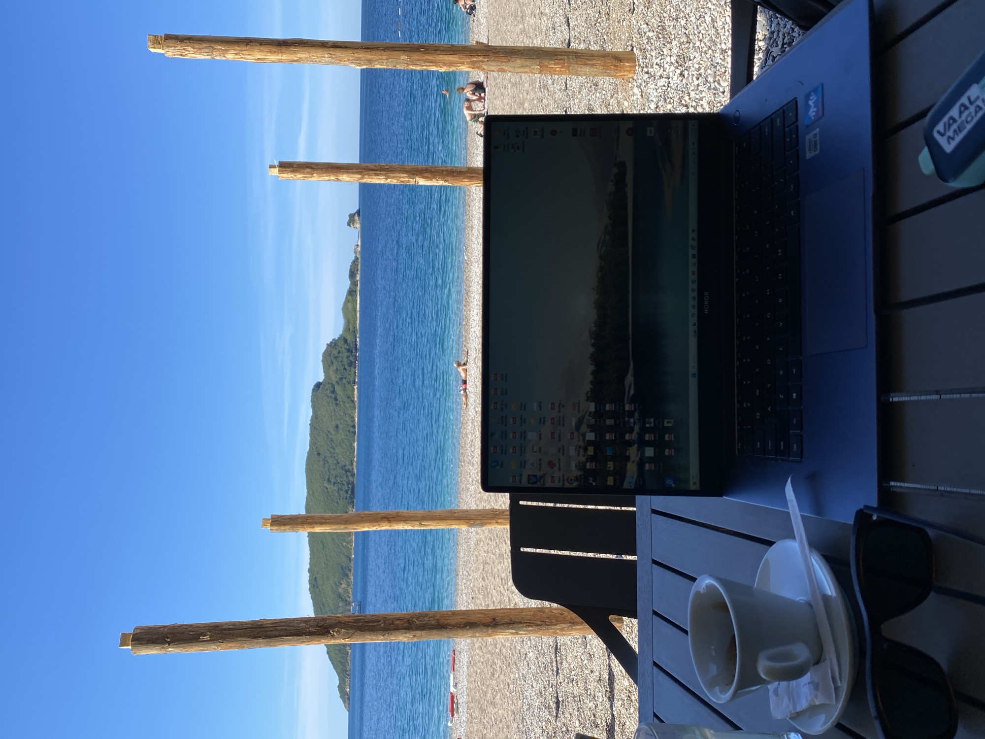 Here's a detailed description of the image:
**Scene:** A workspace on a beach setting, likely at a cafe or outdoor seating area. The beach is in the city of Budva, Montenegro.
**Environment:** The background shows the Adriatic Sea with clear, blue water, a pebble beach, and some rocky cliffs with vegetation. The sky is clear and blue. There are wooden posts that appear to be part of a structure like a sunshade or beach enclosure.
**Objects:**
* A blue laptop is open on the table, partially obscuring the view of the beach.
* A white coffee cup and saucer are on the table, indicating someone has been drinking.
* A pair of sunglasses is on the table.
* A "VAAL MEGA" branded object, possibly a vape or similar item, is also on the table.
* There's a dark-colored chair.
* The table itself is made of wood slats.
**People:**
* There are a few people visible in the background on the beach, some in the water, and some on the sand. They appear to be tourists or beachgoers.
**Actions/Interactions:**
* The scene suggests someone is working or using a laptop while enjoying the beach environment. The coffee cup indicates they've likely been sitting there for a while.
**Overall Impression:** The image portrays a scene of digital nomadism or remote work, where someone is combining work with leisure in a beautiful beach setting.