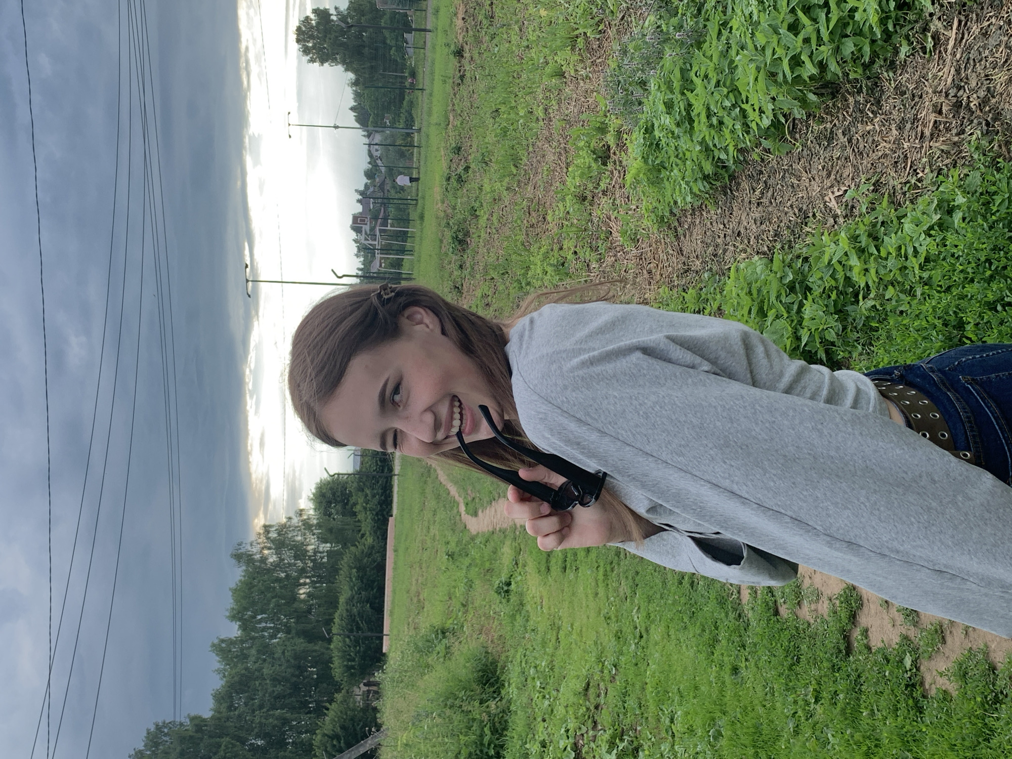 In the city of Fokino, Russia, a young woman with long brown hair, partially pulled back, is captured in a medium shot outdoors. She is looking over her left shoulder towards the camera with a playful expression, one eye closed in a wink and her mouth open in a wide smile. She is holding a pair of black sunglasses with her right hand, playfully biting onto the arm of the glasses. The woman is wearing a light grey long-sleeved shirt and blue jeans, with a dark belt visible around her waist. She is standing on a dirt path that winds through a grassy field. In the background, there are trees, a fence with a soccer goal, and scattered buildings, suggesting a rural or semi-rural setting. The sky is overcast with dramatic clouds, indicating a possibility of rain.