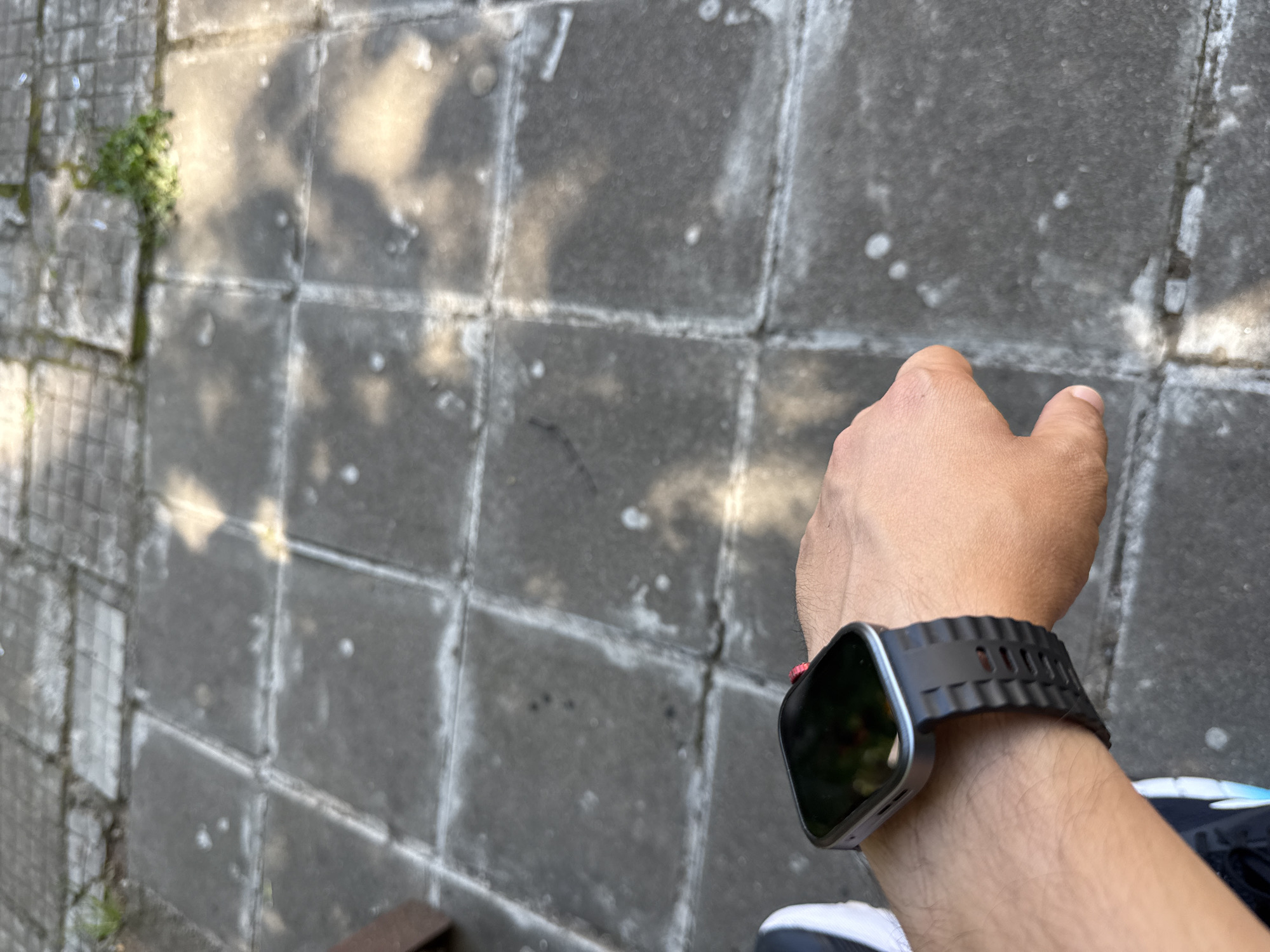 The image shows a close-up view of a person's left wrist, which is adorned with a silver-colored smartwatch with a black, rugged-looking band. The watch face is dark and reflective. The person is wearing athletic shoes, one of which is partially visible in the lower left corner of the frame, featuring a black upper with light blue accents. The background consists of a concrete paved area, likely a sidewalk or patio, with visible square tiles. Sunlight casts shadows on the pavement. There is some sparse greenery growing in the cracks between the tiles in the upper right portion of the image. The scene appears to be outdoors in a city environment. Without further information or context, it is not possible to determine the specific location in Vratsa, Bulgaria, beyond it being an outdoor urban setting.