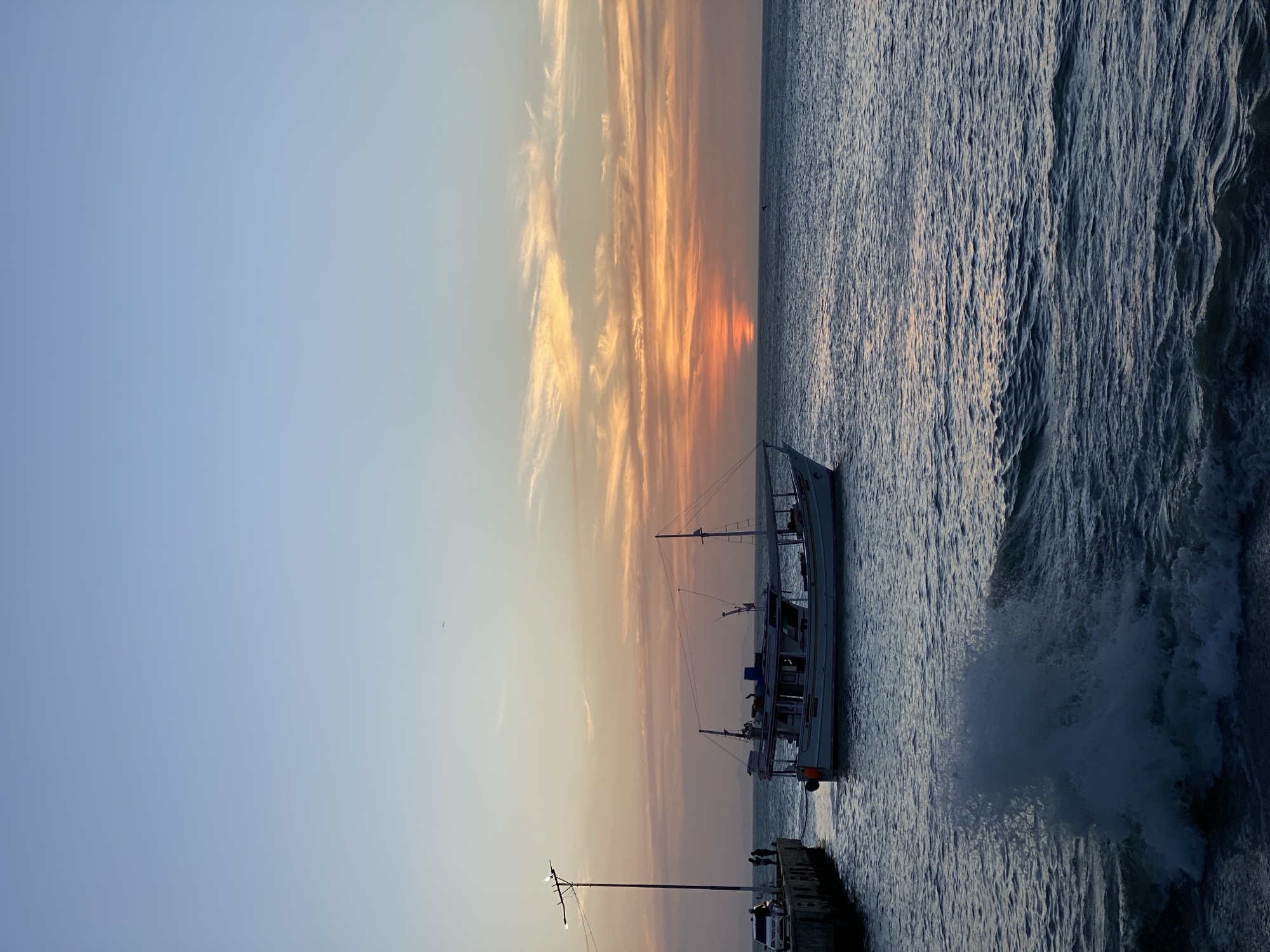 A side view of a ship at sunset is seen in Juan Griego, Venezuela. The sky transitions from clear blue to orange at the horizon. Wispy clouds are scattered above the sun. The sea is rough, with waves crashing against the shore. A pier with a light post and a few people standing on it is visible on the bottom-right of the image. A white car is parked on the pier. The ship is a large fishing vessel. The sun is setting over the sea, creating an orange reflection.