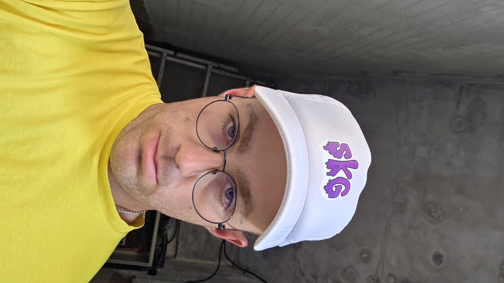 A man wearing a white trucker hat with a purple "SKG" logo, round metal-rimmed glasses, and a yellow t-shirt poses for a selfie in an unfinished concrete interior. The background shows exposed concrete walls and ceiling, a metal ladder leaning against the wall, and some electrical wiring. The lighting suggests an indoor environment, possibly a construction site or unfinished building, in an unknown city.