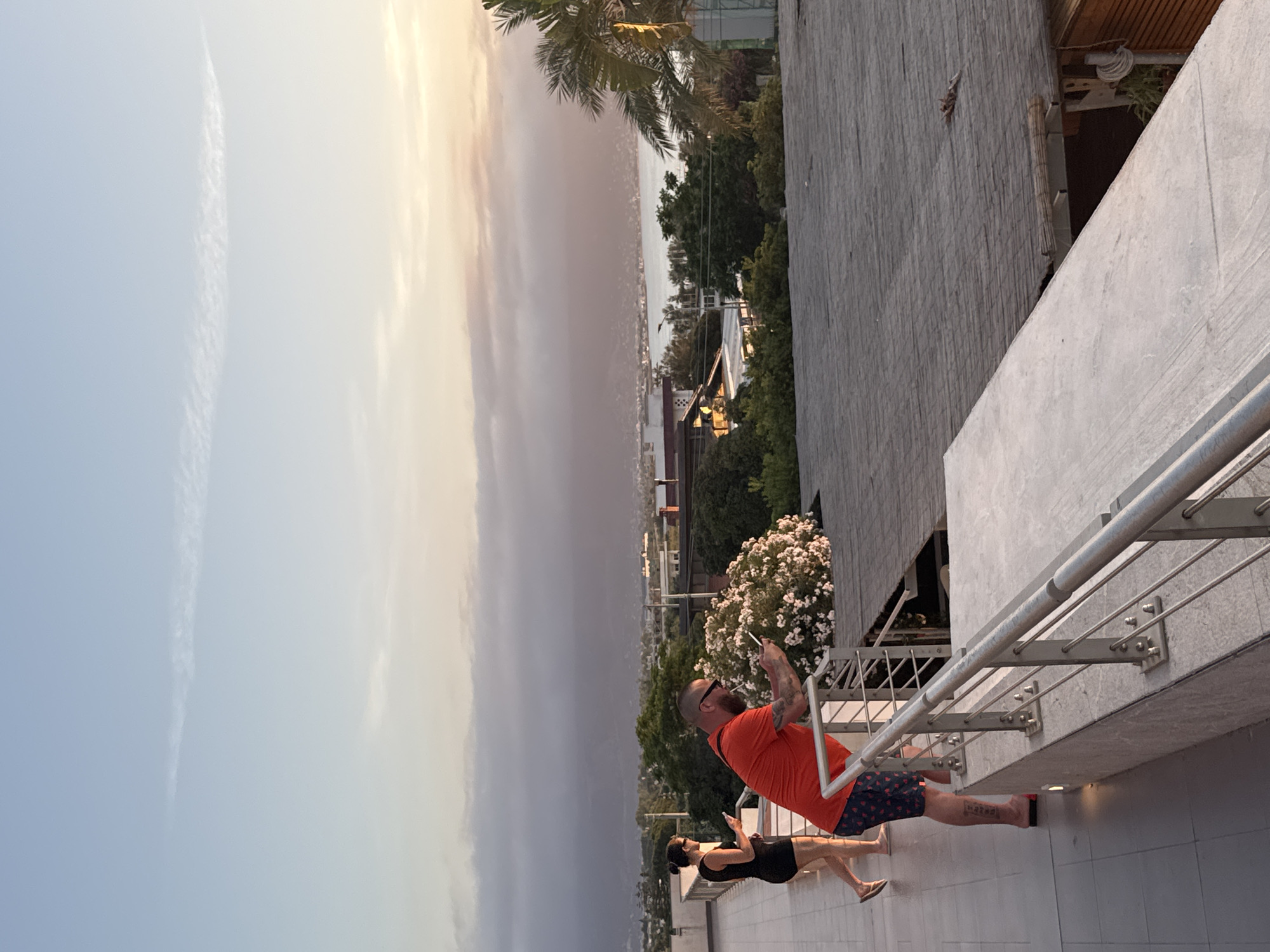 An elevated viewpoint in Isola delle Femmine, Italy, captures a coastal sunset scene. In the foreground, two individuals are on a grey-tiled terrace with a modern metal railing. A man, positioned closer to the camera and leaning on the railing, wears an orange t-shirt, patterned shorts, red flip-flops, sunglasses, and has a beard and calf tattoo; he holds a smartphone and appears to have a white object (possibly a cigarette or vape) in his mouth. Behind him, a woman in a black dress stands upright, also looking at a smartphone. Behind the railing, a large bush with white flowers is prominent. The mid-ground reveals a townscape with various buildings, dense green foliage, and a palm tree on the right. Beyond the town, a bay or sea body reflects the warm light of the setting sun, which casts hues of orange and pink across the horizon. Distant mountains and low-lying clouds obscure parts of the far background, while the upper sky remains a clear light blue with a singular, elongated cirrus cloud. The overall lighting suggests late afternoon or early evening.