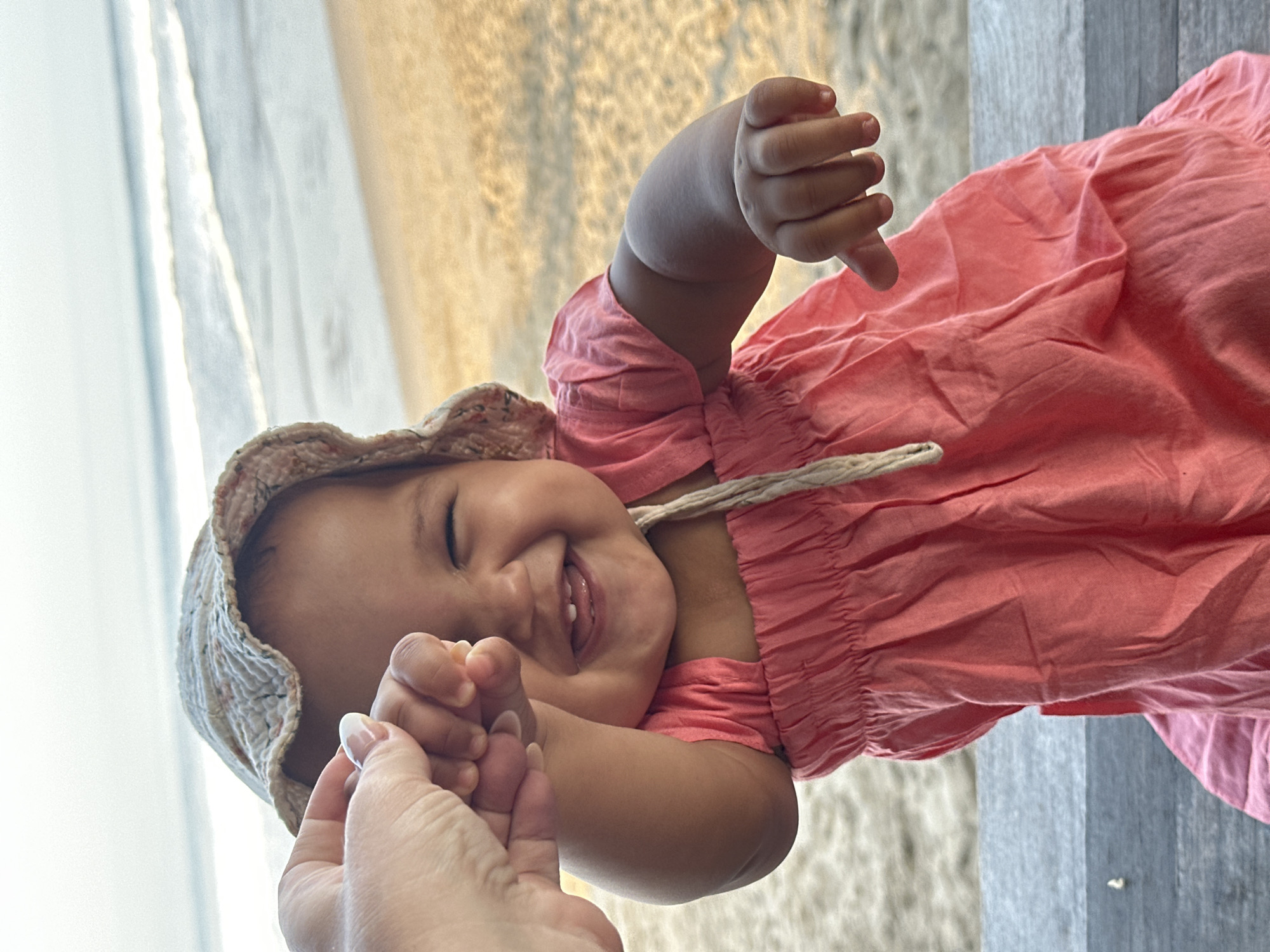A dark-skinned baby wearing a patterned sunhat and a pink dress is held by an adult's hand. The baby is laughing and reaching out with both hands. The scene takes place on a beach in North Kuta, Indonesia, with the ocean and sand visible in the background. The baby is seated on a wooden bench.