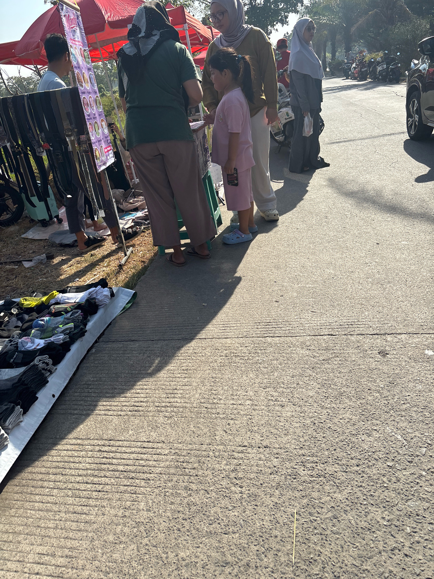 The image captures an outdoor market scene, likely in Telagamurni, Indonesia. In the foreground, a white tarp displays a variety of socks, predominantly black, gray, and white, folded in neat piles. To the left of the socks, a bicycle wheel and part of its frame are visible, suggesting a temporary setup. The ground is paved with concrete, marked by parallel lines, and illuminated by bright sunlight casting long shadows.
In the mid-ground, several people are interacting. A woman wearing a dark hijab and a green t-shirt stands facing away from the camera, near a display banner with colorful circular images, possibly advertising food products. Beside her, a young girl in a pink outfit and light blue clogs stands looking towards the banner. Another woman in a light gray hijab and sunglasses, holding a white plastic bag, stands further to the right. A man in a light blue shirt is visible behind the banner, appearing to be a vendor. In the background, a dark SUV is parked on the asphalt road, and a row of parked motorcycles are visible further down. Palm trees and foliage frame the scene, indicating a warm, sunny environment. The overall atmosphere suggests a bustling local market.