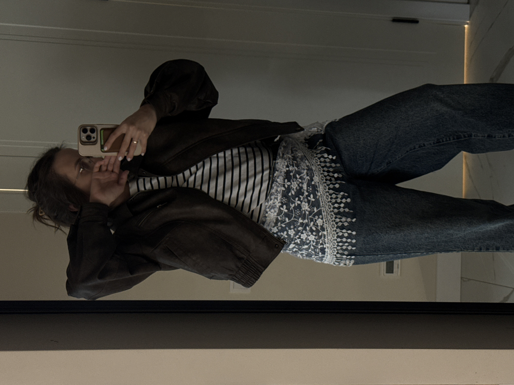 The image displays a mirror selfie of a person, rotated ninety degrees clockwise. The individual, likely female, is shown from the waist up, holding a light-colored smartphone with a visible multi-lens camera array and an attached cardholder. A simple gold-colored ring adorns their left ring finger, and their fingernails are painted with light-colored polish. Their left hand partially covers their lower face.
The person is dressed in a dark brown bomber-style jacket, worn over a white t-shirt with black horizontal stripes. Layered beneath the jacket and over what appears to be blue denim jeans, a blue denim garment with intricate white floral and geometric embroidery is visible, featuring a distinctive patterned hemline.
The background, reflected in the mirror, consists of light-colored interior walls or doors, with one featuring a dark horizontal handle. A subtle warm vertical light strip is visible on the right side of the reflection. The scene is clearly indoors. No specific time of day or weather conditions are discernible. There is no visible text within the image. The location context provided is Pervomaysky, Russia.