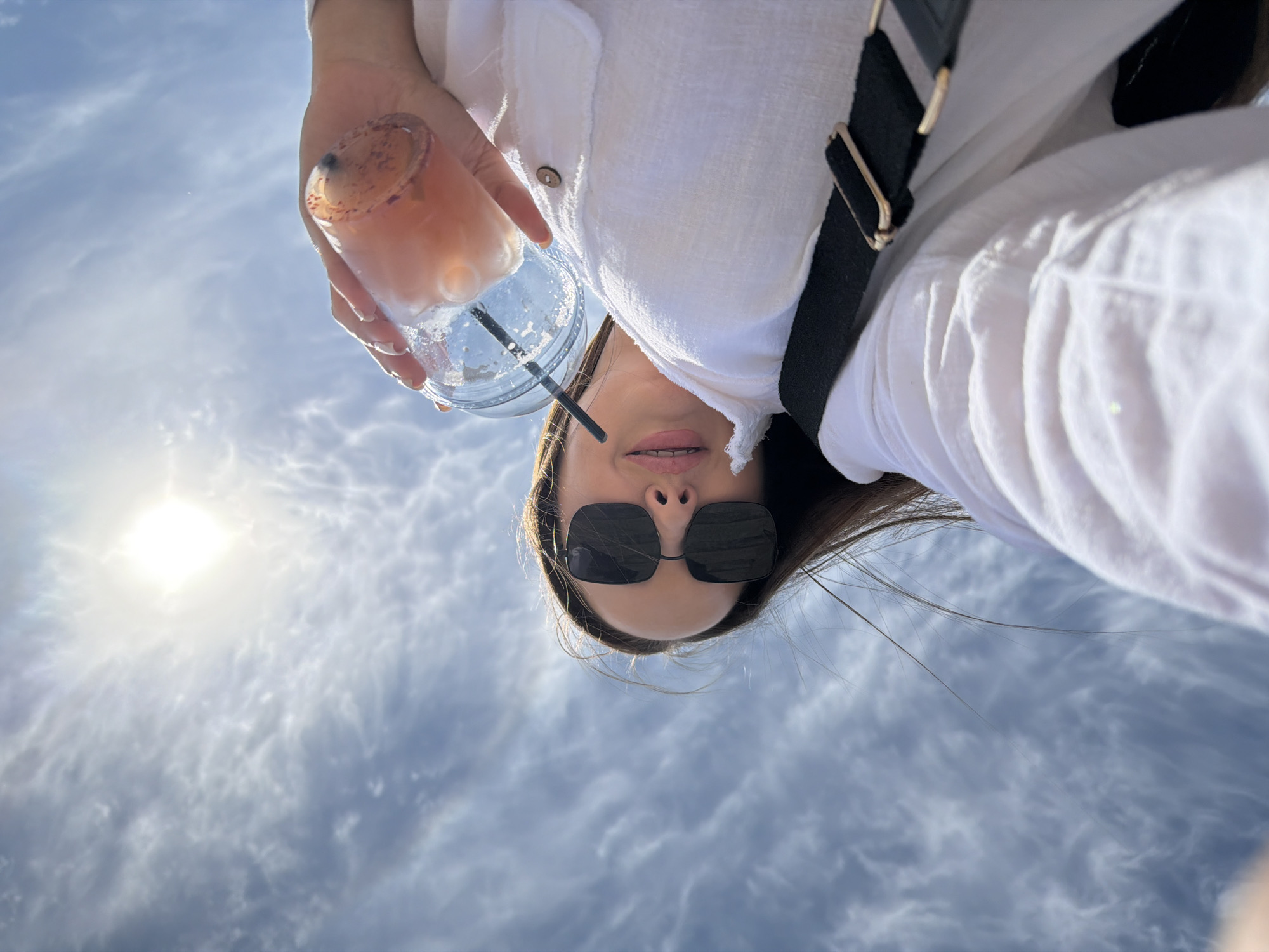 A low-angle shot depicts a woman wearing sunglasses and a white linen shirt. She holds a clear plastic cup with a straw, containing a light-colored beverage. A black bag strap is visible across her shoulder. The background features a cloudy sky with the sun shining brightly. The location is not evident from the image.