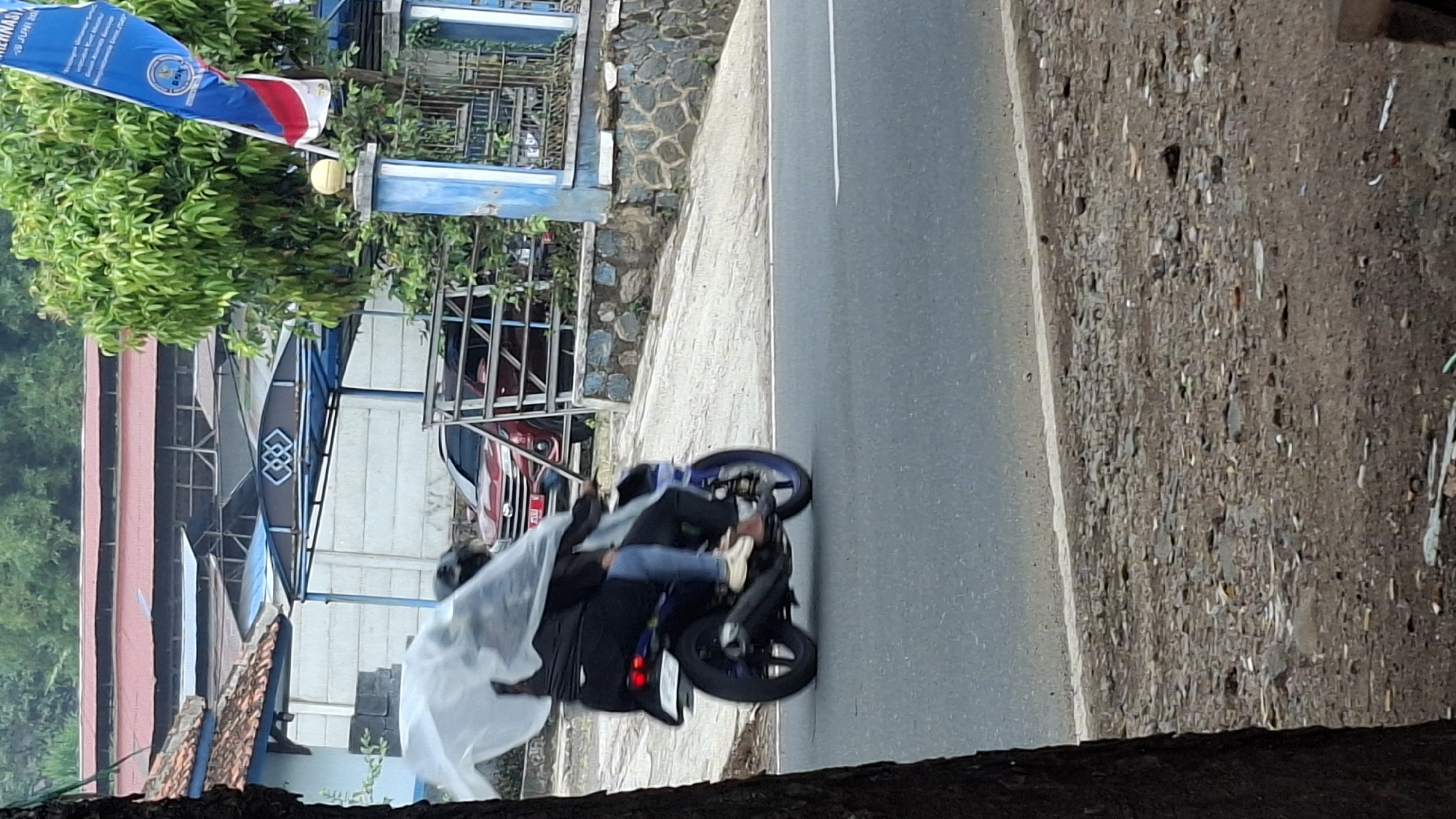 In Cilaku, Indonesia, a motorcyclist and a passenger are traveling on a street. The motorcyclist is wearing black clothing and blue jeans, with white shoes. The passenger is also wearing black clothing. They are both covered by a clear plastic sheet, possibly to shield them from rain. The motorcycle is blue and black with blue rims. The street is paved, and there is a stone wall and some vegetation on the right side of the road. In the background, a red car is visible, as well as buildings with red roofs. A flag with blue and red colors is also seen. The environment appears to be an outdoor setting in a town or city.