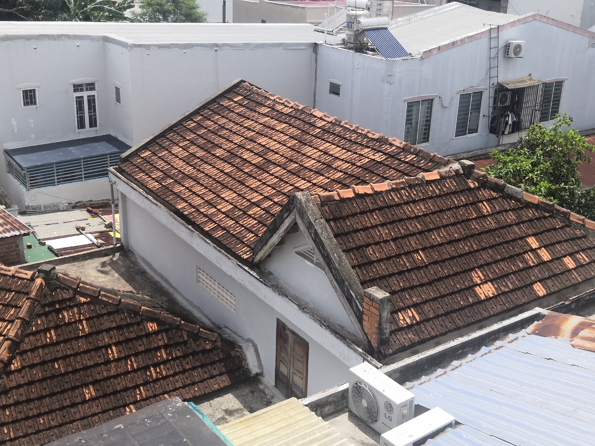 The image shows an aerial view of rooftops in a densely populated urban area, likely Bac Nha Trang Ward, Vietnam. The dominant features are terracotta tiled roofs, some appearing weathered and aged, interspersed with corrugated metal roofing and flat, white-plastered buildings. Several buildings have visible air conditioning units mounted on their exteriors. One building features a balcony with a metal railing and a dark blue flooring. Another building has a ladder extending up its side, and laundry is visible hanging from a window. The overall impression is one of a bustling city with residential and commercial structures closely packed together. There are no people visible in the image. The environment is characterized by clear daylight.