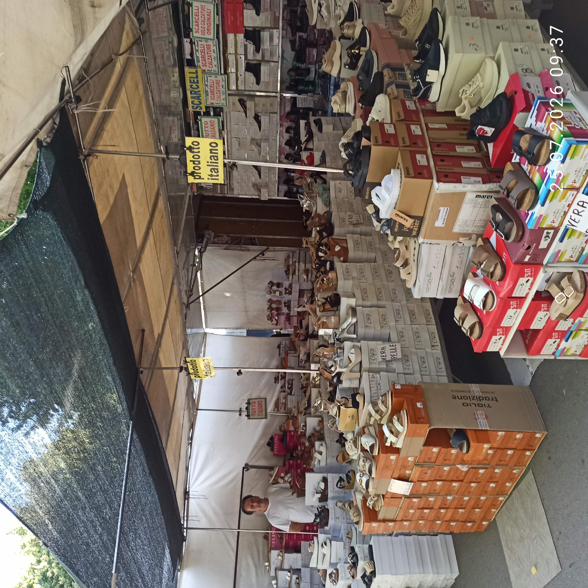 A man in a white t-shirt is standing behind a stall selling shoes in Ruvo di Puglia, Italy. The stall is covered by a dark green mesh tarp and displays a variety of footwear, including sandals and sneakers, organized in boxes and on shelves. Yellow signs with Italian text, such as "prodotto italiano" (Italian product) and "SCARCELLI CALZATURE" (Scarcelli Footwear), are visible, indicating the origin and type of goods. The man appears to be attending to customers, though none are directly interacting with him in this frame. The environment suggests an outdoor market or fair, with a paved ground and other stalls visible in the background. The image is dated "25.07.2026 09:37".