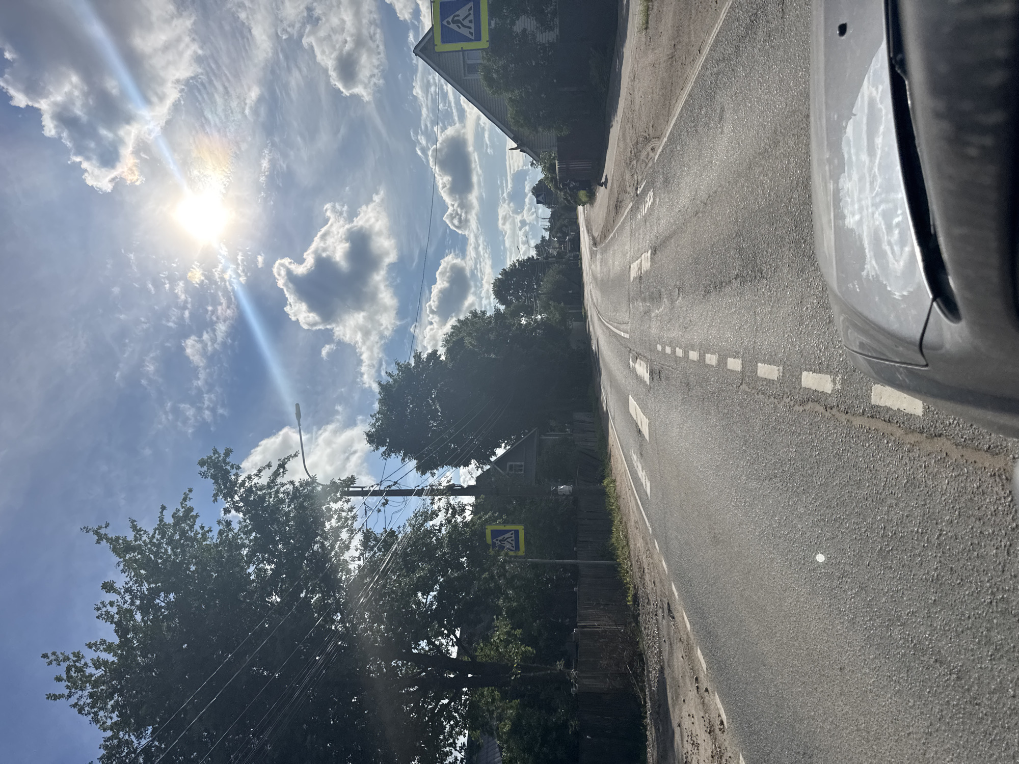 The image depicts a sun-drenched road in a rural or suburban setting, possibly Saint Petersburg, Russia. The sun is high in the sky, casting bright light and lens flares through a partly cloudy sky, suggesting it is daytime, likely midday or early afternoon. The weather appears warm and pleasant.
The scene focuses on a paved road with white dashed lines marking the lanes, indicating it is a public road. On the right side of the road, a portion of a car is visible, suggesting the image was taken from inside a vehicle. The road is bordered by trees and some houses, giving a sense of a community. There are two pedestrian crossing signs visible, one on the left and one on the right side of the road, warning drivers of potential pedestrian crossings. Power lines and a utility pole are also present, adding to the infrastructure detail.
The overall mood is one of a bright, ordinary day. There are no people clearly visible in the image, and the focus is on the landscape and the road itself. The scene is characterized by natural elements like the sun, clouds, and trees, juxtaposed with man-made structures like the road, signs, and power lines. The presence of the car suggests movement or travel, but no specific activity or event is actively taking place within the frame. The image conveys a sense of peacefulness and the simple beauty of a sunny day.