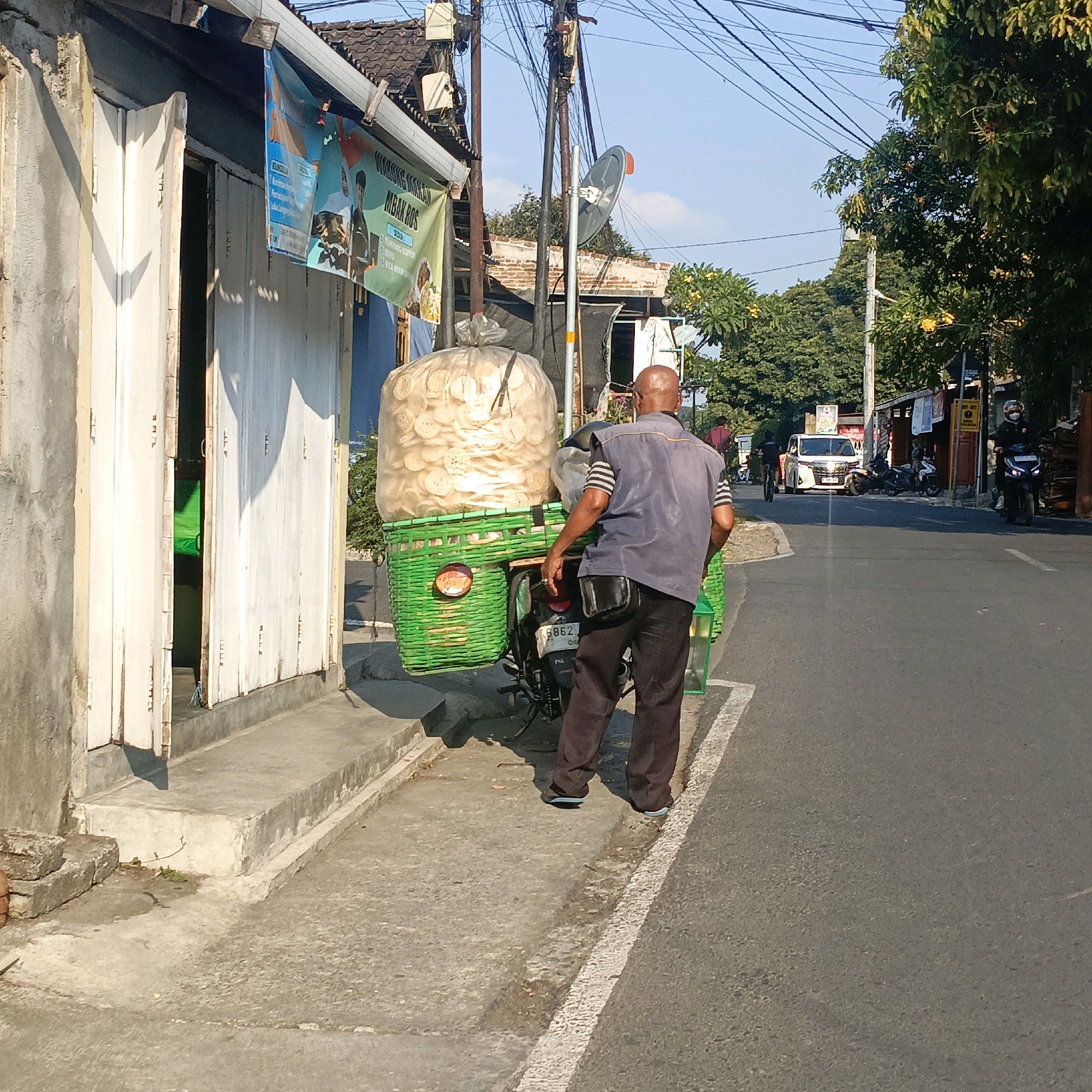 An Indonesian man, bald and wearing a dark blue vest over a striped shirt and dark pants, stands on a sidewalk next to a motorbike loaded with a large green basket filled with a huge bag of light-colored, circular snacks, possibly crackers or crisps. The motorbike's license plate is partially visible, reading "B 3862 OS". The man is facing away from the camera, towards a street where a white van is driving past. The scene appears to be in Salatiga, Indonesia, indicated by the street signs and architecture. The building to the left has a white-paneled door and a banner with Indonesian text that reads, "WARUNG MAKAN MBAK ROS". The street is paved with asphalt, and utility poles with numerous wires are visible against a blue sky. Trees line the street, and other vehicles, including motorcycles, are present in the background. The overall impression is of a typical street scene in an Indonesian city, likely during daylight hours.