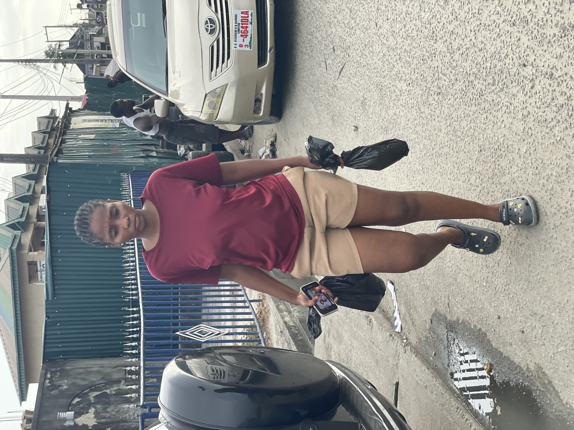 An upright, eye-level shot captures a young woman standing on a paved street in what appears to be an urban setting, identified as Ado, Nigeria. She is positioned slightly to the left of the frame's center, looking directly at the viewer with a neutral expression. She wears a maroon t-shirt, light brown shorts, and dark Crocs adorned with gold charms, some showing the text "New Brilliant." In her left hand, she holds a smartphone displaying an image, and in her right hand, she carries two black plastic bags.
Behind her, partially visible in the mid-ground, is a dark-colored car. Further into the background, a white Toyota car is parked, its front license plate clearly visible, reading "F.A. ELEGUSHI S.O. GLOBAL LIMITED" and "03 - 4641DLA FEDERAL REPUBLIC OF NIGERIA." Next to the white car, a man in a white tank top and dark shorts stands, looking towards the left. Another person's arm is faintly visible behind the white car.
The background features a mix of buildings, including structures with green corrugated metal walls and another with a green roof, indicating a bustling, built-up area. A blue metal fence with decorative spikes runs across the mid-ground. Overhead, numerous electrical wires crisscross between utility poles against a bright, overcast sky, suggesting daytime.
The street surface is light grey asphalt or concrete, showing some wear. In the bottom right corner, a puddle of water with a visible metal drain grate reflects the sky. Scattered debris, including pieces of paper, lies on the ground near the puddle. The overall atmosphere suggests a typical day on a city street.