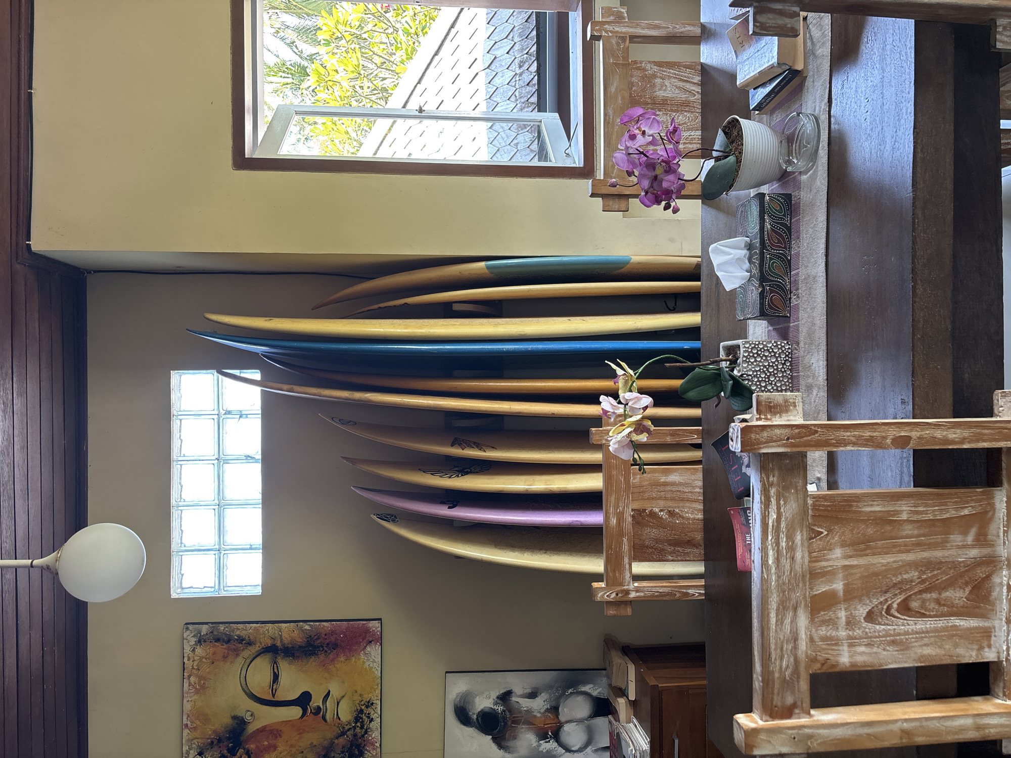 The interior of a building in Pekutatan, Indonesia, is shown. A collection of surfboards is mounted horizontally on a beige wall. Underneath, two framed pieces of art hang. A white globe lamp is on the left. A window above the surfboards lets in natural light. To the right, a wooden table and chairs occupy the space. Floral arrangements and decorative objects are placed on the table, near a stack of books.