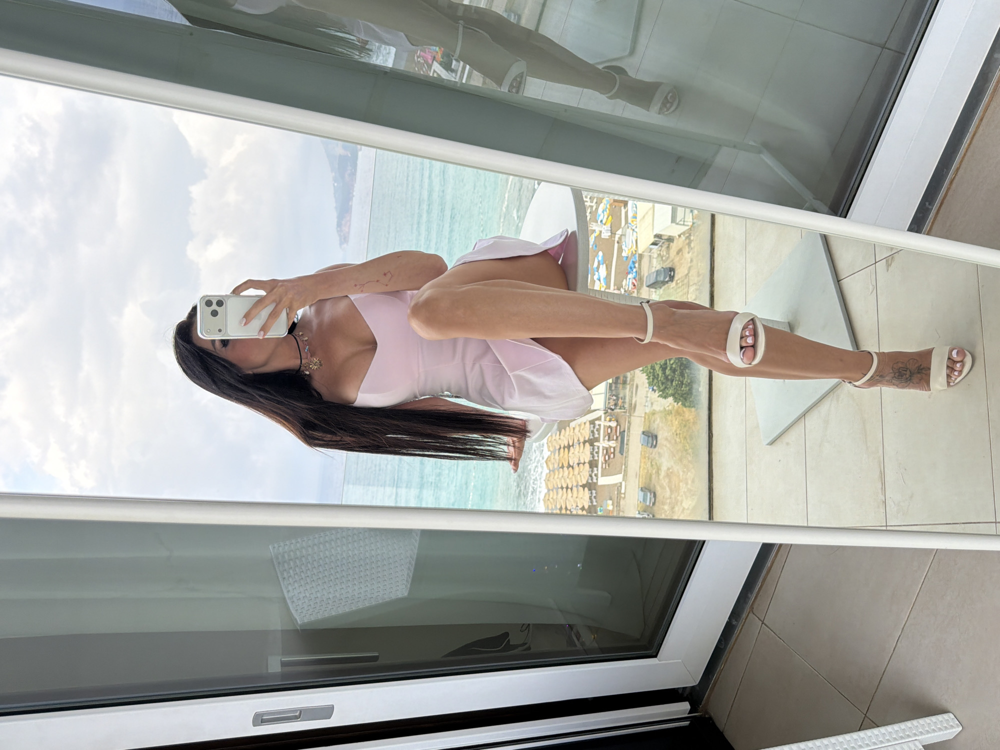 A woman is captured in a mirror selfie taken on a balcony overlooking a coastal landscape. She stands, reflected in the large glass panel, wearing a light pink mini-dress and white heeled sandals. Her long, dark hair cascades down her back. She holds a white smartphone with multiple cameras up to take the photo, partially obscuring her face. Visible accessories include a necklace with a shell-like pendant and a white bracelet on her left wrist. A floral tattoo adorns her right foot/ankle, and her fingernails and toenails are painted a light pink.
The mirror reflects a vibrant outdoor scene. A calm, turquoise sea stretches out under a partly cloudy sky. Along the shore, a sandy beach is densely covered with rows of straw umbrellas and sun loungers, suggesting a busy resort area. Beyond the beach, some buildings and a road with vehicles are visible. The balcony itself features a light-colored tiled floor. The setting suggests a daytime scene, possibly in Boreti, Montenegro, with diffused natural light.