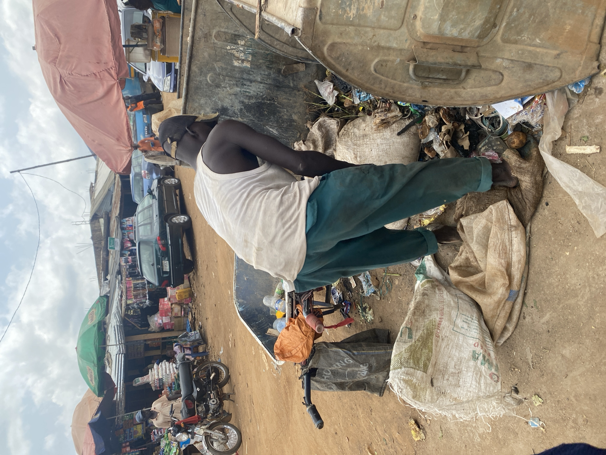 The image shows a man in a white tank top and green pants standing beside an overflowing trash can in what appears to be a market or street scene in Kafanchan B, Nigeria. The trash can is filled with various types of refuse. The man is sorting through trash. There are several bags lying on the ground and a wheelbarrow filled with refuse and plastic bottles. The background includes various shops, motorcycles, cars, and umbrellas, suggesting a bustling urban environment.