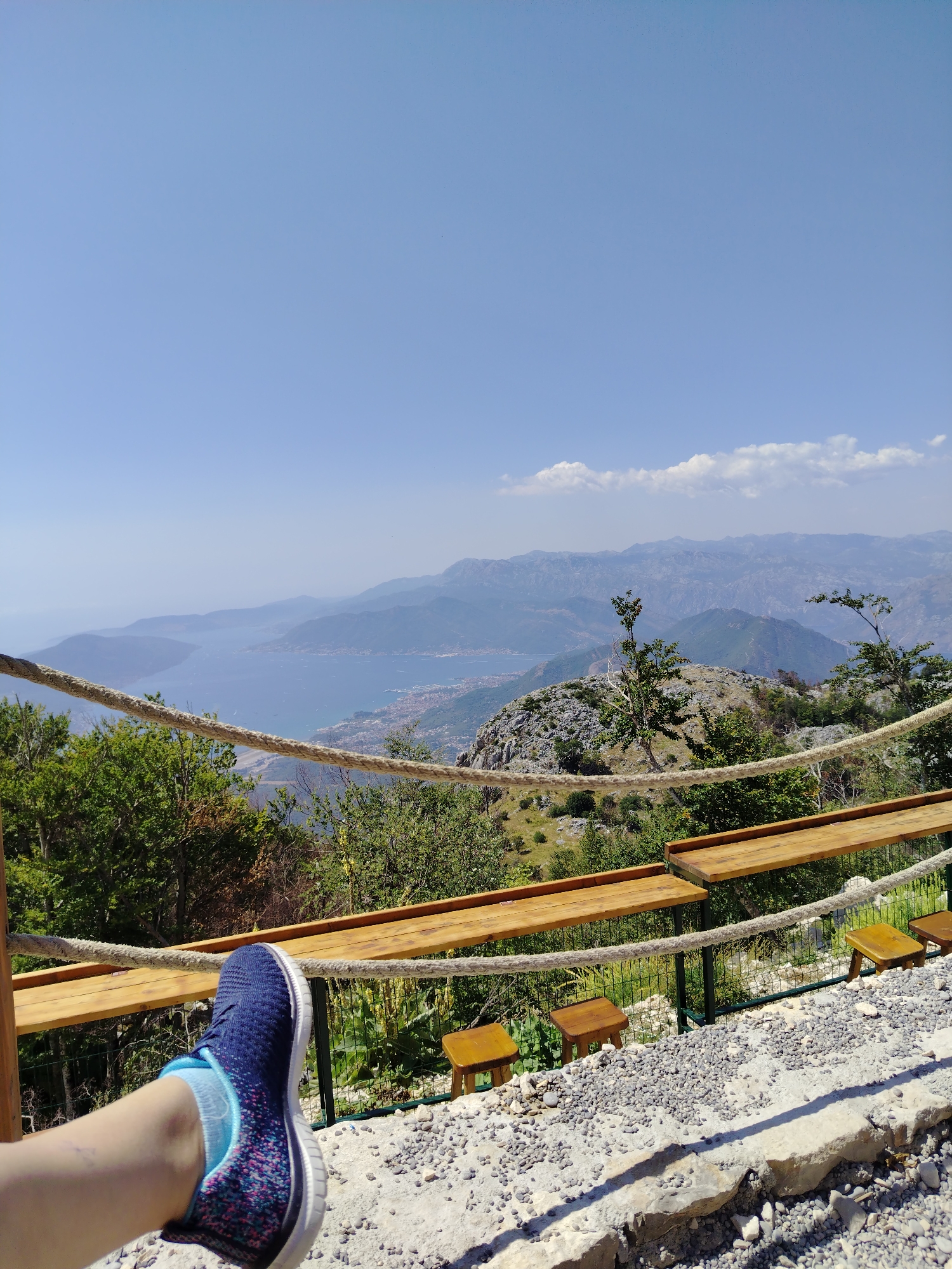 The image depicts a scenic viewpoint overlooking a bay and mountains, likely situated in the vicinity of Kuk, Montenegro. In the foreground, a person's left leg is visible, clad in a blue and pink speckled sneaker and light blue socks. The leg is positioned in a relaxed manner, resting on a stone and gravel surface. A thick rope forms a barrier in front of the viewer, suggesting a safety railing for the overlook.
Beyond the rope, a wooden bench and small stools are arranged along the edge of the viewpoint, providing seating for visitors. The landscape unfolds with lush green trees and rocky terrain leading down to a wide expanse of blue water, which appears to be a bay or gulf. In the distance, a city or town is visible along the coastline, with a backdrop of layered, hazy mountains that fade into the sky. The sky itself is a clear, pale blue with a few wispy clouds on the horizon. The overall impression is one of a serene and picturesque natural setting, likely a popular tourist destination for enjoying panoramic views.