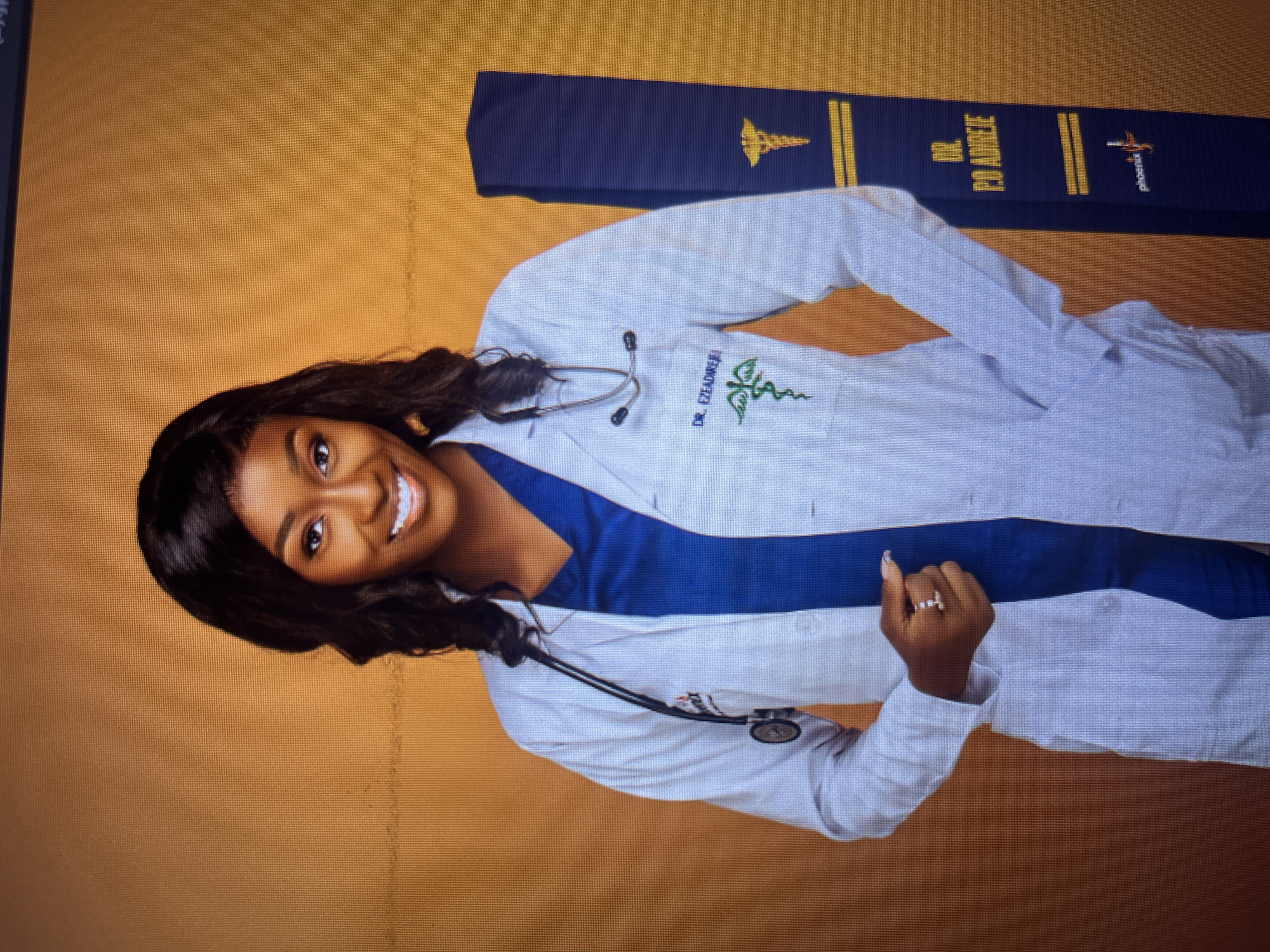 A medium shot shows a Black woman, who appears to be a doctor, standing against a textured orange background. She is wearing a white lab coat over a royal blue V-neck scrub top. A stethoscope is draped around her neck. Her lab coat has an embroidered caduceus symbol in green and the name "DR. EZEADIREREI" embroidered in blue on the left chest. She is smiling and looking at the camera. Her left hand, adorned with a ring featuring three clear stones, is resting in the pocket of her lab coat. To her right, a dark blue sash with gold lettering and symbols is leaning against the background. The sash has "DR. P.O. ADIREJE" embroidered in gold letters, along with a caduceus symbol and a stylized star logo that reads "phoenix". The overall scene suggests a graduation or professional portrait. There is no visible indication of the location within the city of Gura Top, Nigeria.