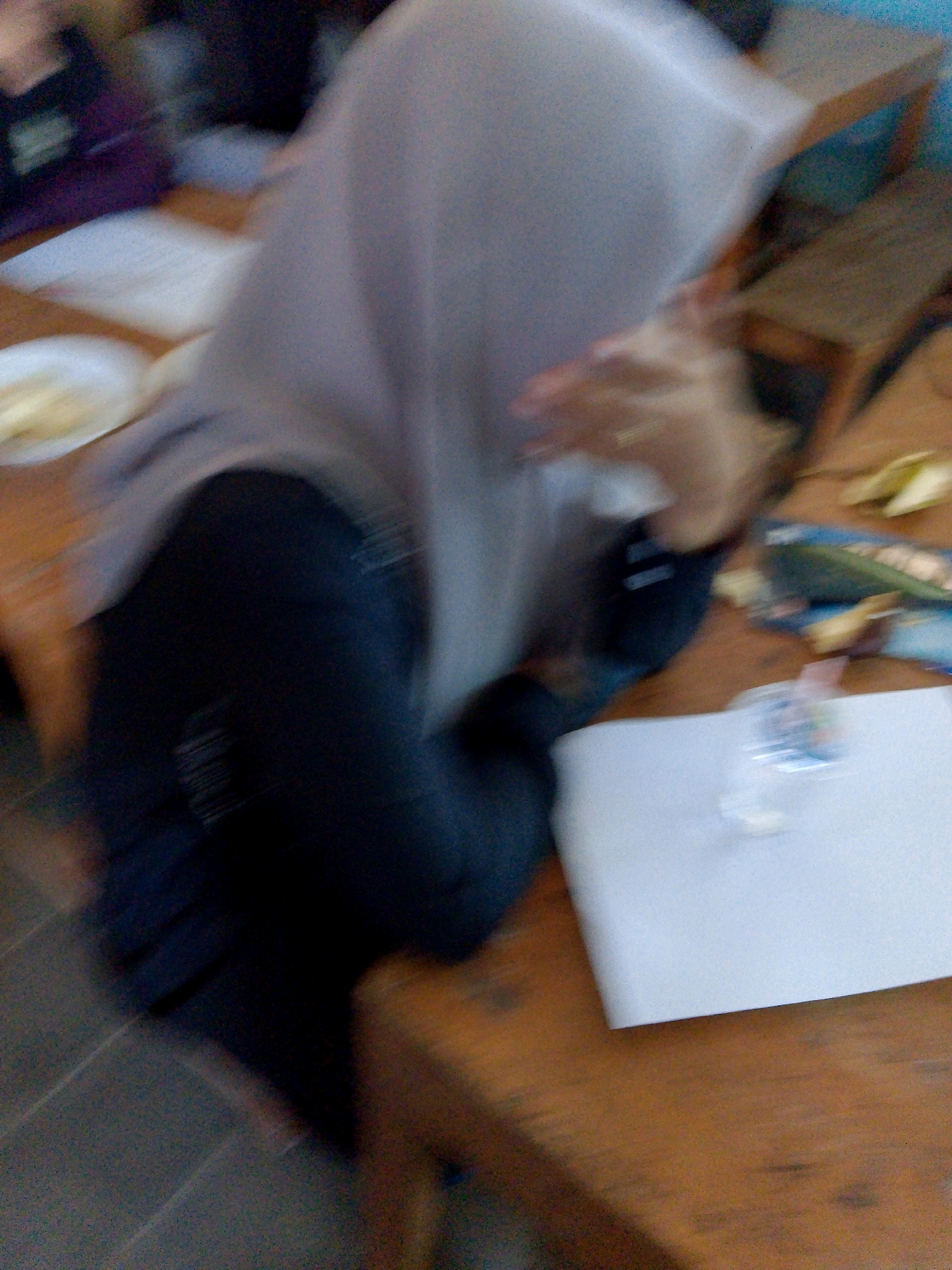 The image depicts a blurred scene on a wooden table, likely in a classroom or dining area in Kuripan Utara, Indonesia. A person wearing a light-colored hijab and a dark-colored garment is partially visible, with their right hand near their face. On the table, there are a white plate with some food, a notebook, and a small clear container with a colored liquid. The background is also out of focus, showing hints of other people and furniture.