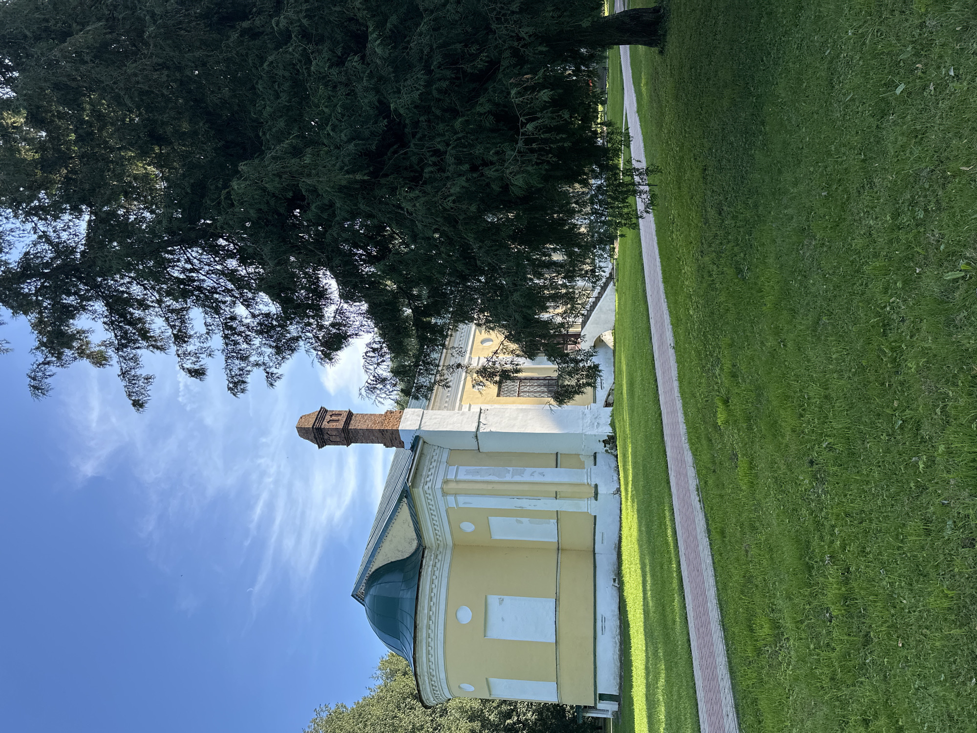 The image depicts a section of the grounds surrounding the historical building of the Selivanovo Glass Factory Museum in Selivanovo, Russia. The primary subject is a pale yellow, oblong building with a green, domed roof. A prominent red brick chimney stands next to the building, which also features white decorative elements and boarded-up windows. The building is situated on a well-maintained lawn. A red and white brick pathway curves through the grass, leading towards the building. To the right of the pathway, a large, mature evergreen tree with dense foliage casts a shadow across the scene. The sky above is blue with scattered wispy clouds. The overall impression is one of a serene, historical park setting on a clear day.