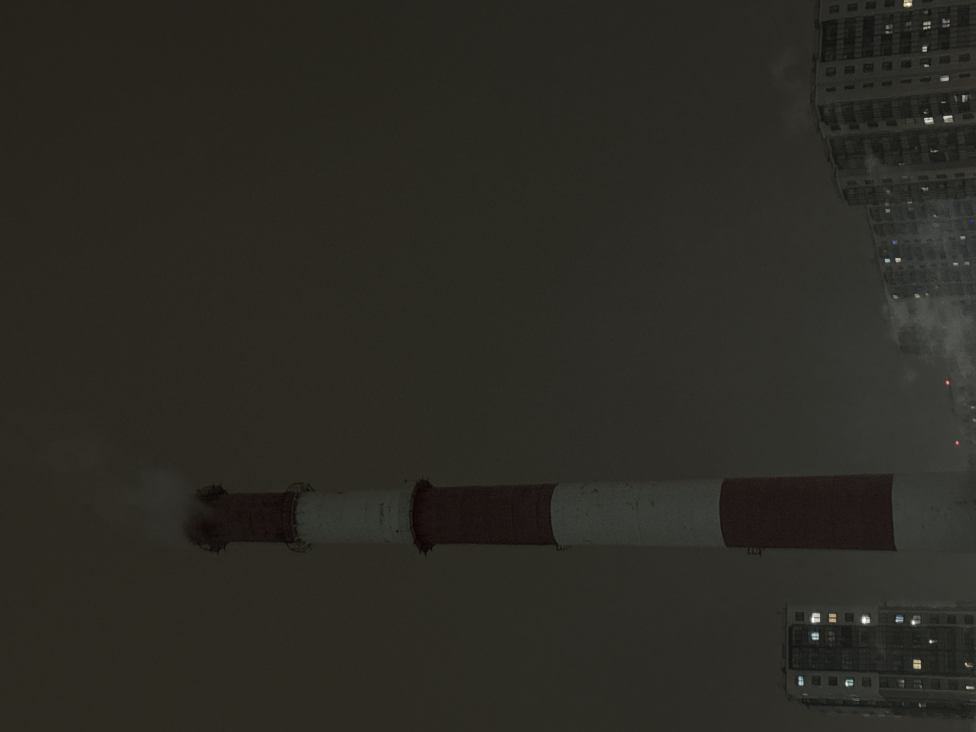 The image presents a nocturnal urban or industrial scene in Saint Petersburg, Russia, captured from an unusual, possibly upward-tilted perspective. The primary subject is a large industrial chimney, oriented horizontally across the frame, characterized by distinct red and white horizontal stripes. From its left end, a visible plume of white smoke or steam is emitted, dissipating into the dark, featureless sky.
On the right side of the frame, and vertically aligned, are multiple sections of what appear to be tall residential or office buildings. Their windows are illuminated with various bright lights, suggesting interior activity or habitation. Another portion of a building is visible at the bottom of the frame, also showing illuminated windows. The sky is uniformly dark gray, indicating nighttime or heavily overcast, possibly foggy, conditions, which contribute to a subdued and stark atmosphere. No individuals or vehicles are discernible in the scene. There is no visible text present in the image.