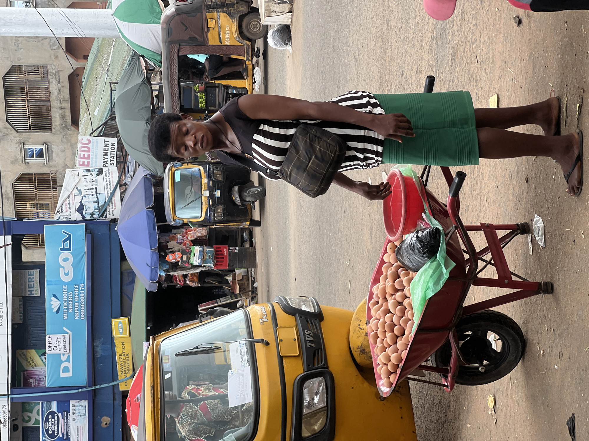 A woman stands on a street in Nkpor, Nigeria, next to a wheelbarrow filled with eggs. She is wearing a black and white striped top and a green skirt. A dark shoulder bag is slung across her chest. The wheelbarrow is red and has two wheels. Behind her, a yellow tricycle-like vehicle is parked. In the background, various buildings and signs are visible, including one that reads "GOtv" and another for "EEDC PAYMENT STATION". Several other vehicles and street vendors are also present in the scene. The ground is paved with asphalt and appears somewhat dusty. The lighting suggests it is daytime.