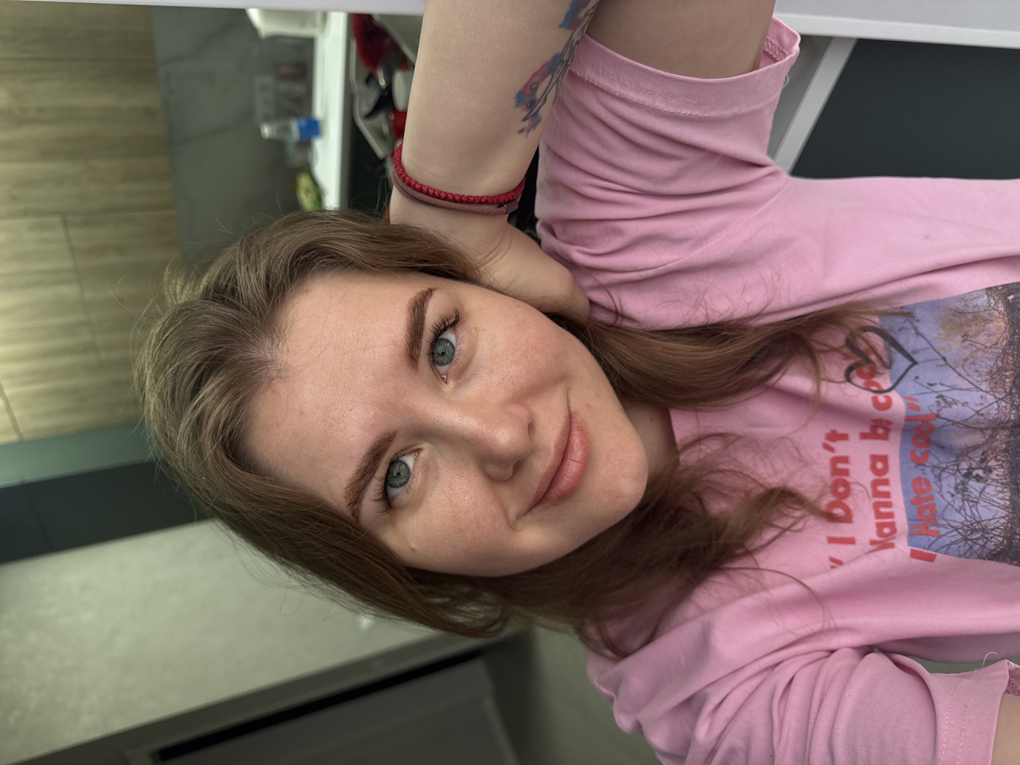 A young woman is photographed at a close range, likely in an indoor setting. The individual has light brown hair, blue eyes, and fair skin. She is wearing a pink shirt with a printed design and text on the front. A red bracelet adorns her left wrist, and a tattoo is visible on her arm. The background is a blurred combination of light-colored surfaces, including what appears to be a wall, shelf and a bathroom-like area. The photograph provides no information to identify a specific location within Gornyi.