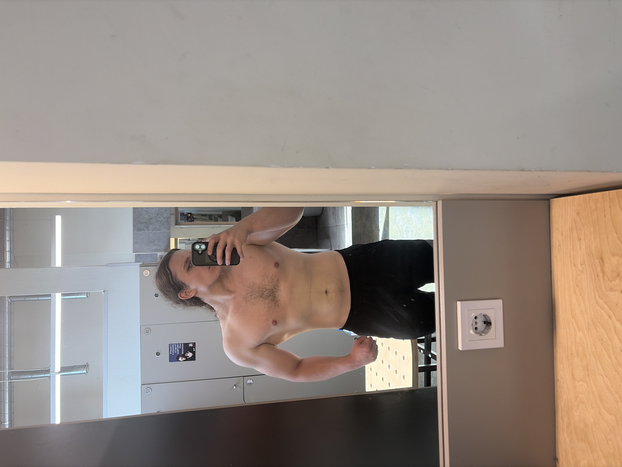 A shirtless man is taking a selfie in a mirror. He is wearing black pants. The reflection shows lockers and a radiator in a room. An electrical outlet is visible next to the mirror. The location is likely a gym or changing room in Plovdiv, Bulgaria.