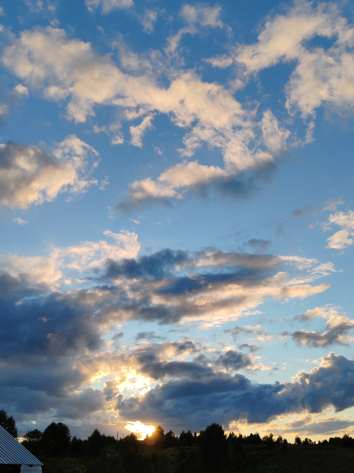 The image depicts a twilight sky with dramatic cloud formations over a silhouetted landscape. The sky is a gradient of blues, with streaks of warm, golden light breaking through the dense clouds, suggesting the sun is setting or has recently set. Scattered white and gray clouds are present throughout the frame, some appearing wispy and others more substantial. The foreground consists of a dark, tree-lined horizon. A portion of a white, corrugated metal roof of a structure is visible in the lower left corner. The overall impression is one of a rural or natural setting in the city of Чухлэм, Russia, during the transitional period of dusk.