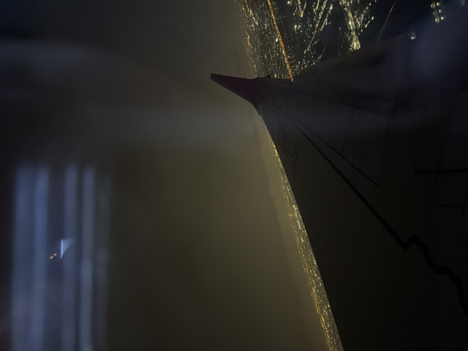 An aerial view at night captures a portion of an aircraft's dark grey wing, prominently positioned in the middle ground, extending from the lower left towards the upper right. The wing displays faint markings, including "D-HHK," and features a small section of a purple or dark red wingtip. Below the wing and across the horizon, the urban sprawl of Racha Thewa Subdistrict, Thailand, is illuminated by a dense grid of warm-toned city lights. The sky above is dark and hazy, with a greenish-brown tint visible near the horizon. A distinct, bright vertical beam of light, originating from an unseen source, cuts through the atmospheric haze in the upper-middle section of the frame. Reflections of interior cabin lights or structural elements are present as faint horizontal streaks and a small luminous point in the upper left corner of the image.