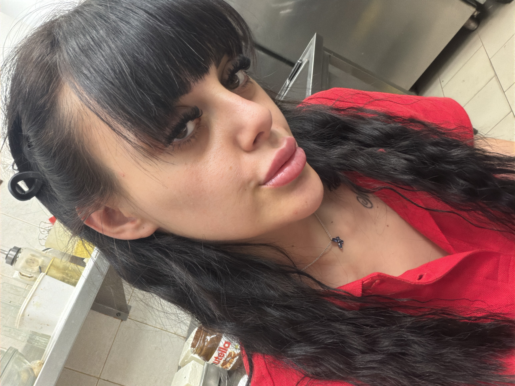 A close-up, selfie-style shot captures a young woman with long, dark, wavy hair and bangs. She is wearing a bright red collared shirt. A delicate silver necklace with a dark blue pendant is visible around her neck. Her makeup includes prominent false eyelashes and lip gloss.
The background suggests an indoor setting, possibly a kitchen or restaurant. A stainless steel shelf is visible to the left, holding various items including a glass dispenser with a metal cap, a white bucket, and a jar of Nutella. The tiled walls and floor further indicate a commercial or food-service environment. A hint of an appliance, perhaps an oven or freezer, is visible in the upper right portion of the frame. The lighting appears to be artificial, consistent with an indoor location. No specific time of day or weather conditions can be determined. The woman's expression is relaxed, with her head tilted and looking towards the camera. The overall impression is informal, possibly a moment captured during a break.
Visible text: "Nutella" is clearly legible on the jar in the background.