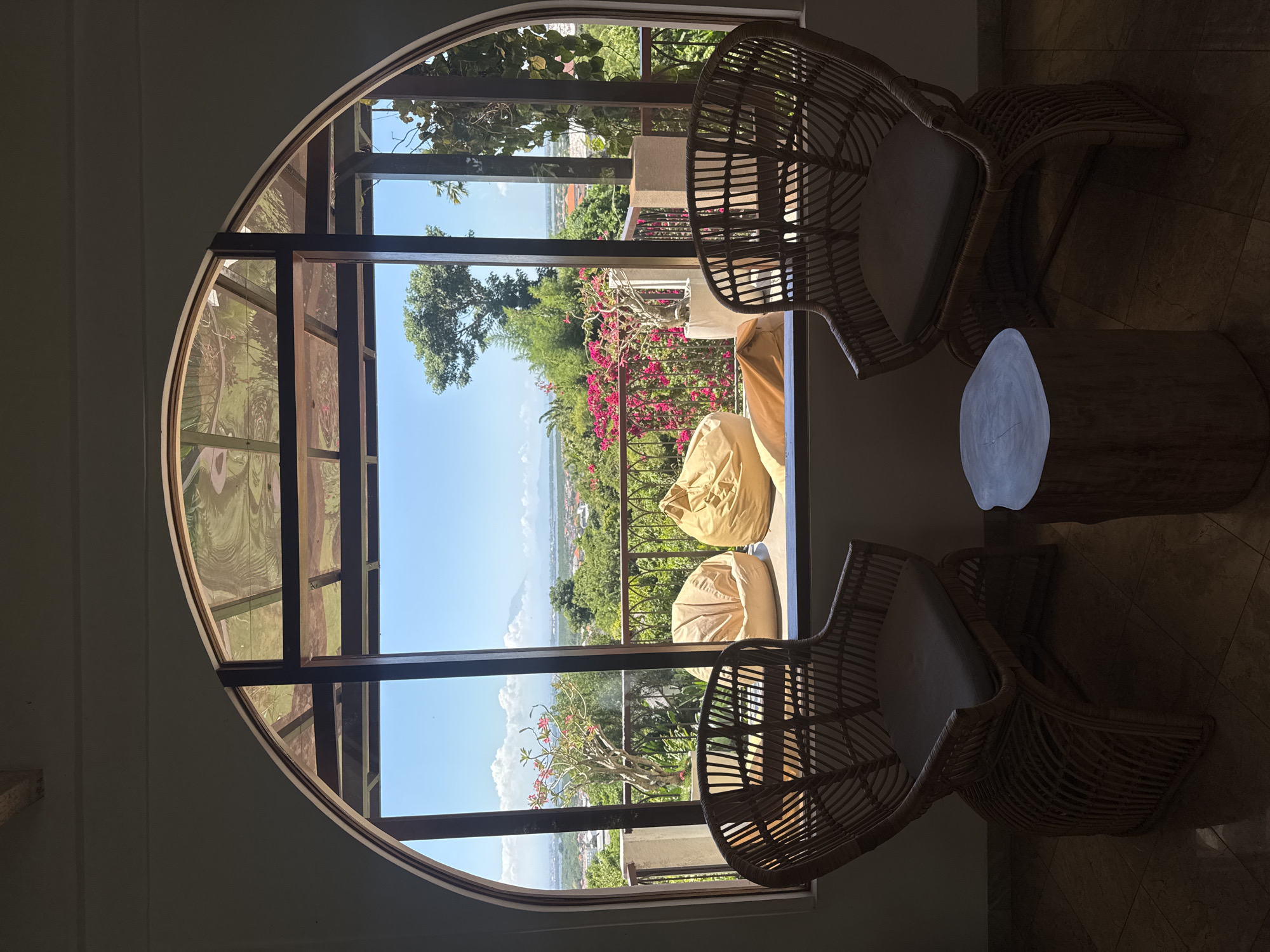 The image shows an interior view through a large, arched window overlooking the city of Kuta Selatan, Indonesia. The window is divided into rectangular panes by dark wooden frames. Outside the window, a balcony is visible with pink flowers and two tan-colored beanbag chairs. Beyond the balcony, the landscape slopes down towards the ocean, with trees, buildings, and a clear blue sky.
Inside, two rattan chairs with light cushions are positioned near the window, facing towards the view. A small, round wooden table stands between them. The lighting is dim in the foreground, contrasting with the bright outdoor scene. The room's walls appear to be a light neutral color. The scene suggests a relaxed or contemplative space oriented towards the outdoor scenery.