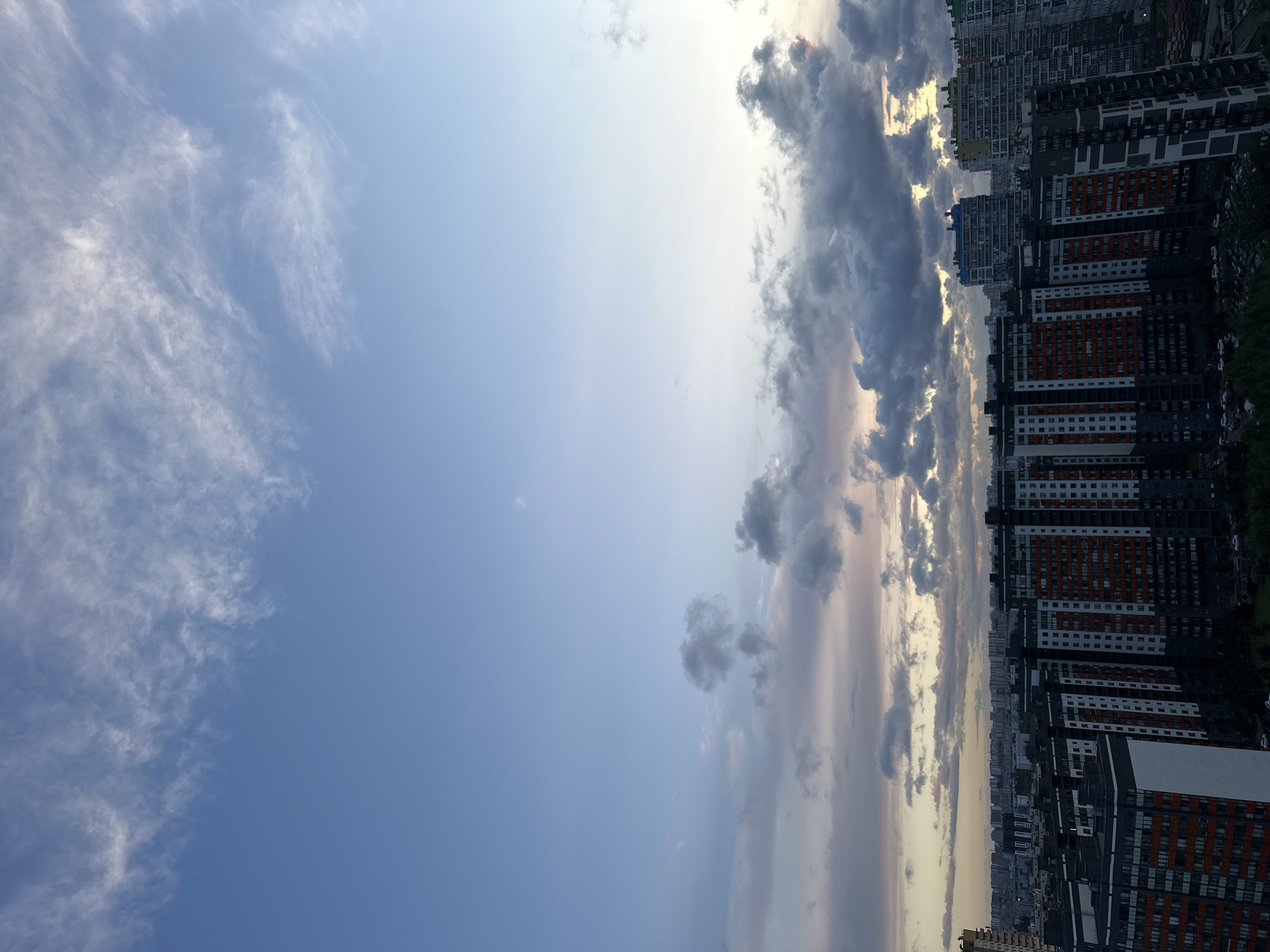 This image, taken from a high vantage point, shows a sprawling cityscape with a dramatic sky. The foreground is dominated by a dense cluster of modern high-rise apartment buildings. These buildings feature a mix of dark gray, white, and orange facades, with many balconies visible. The architecture is consistent with contemporary urban development, suggesting a recently built or rapidly developing area.
In the middle ground, more apartment buildings of varying heights and architectural styles stretch across the horizon. Some are lighter in color, with windows reflecting the ambient light. Further in the distance, a vast expanse of lower-rise buildings forms a dense urban fabric, characteristic of a large city.
The sky above is a prominent feature. The upper portion is a clear, pale blue, transitioning to softer hues of pink and yellow closer to the horizon. Wispy, thin clouds are scattered across the upper sky, while thicker, darker clouds gather lower down, creating a sense of depth and atmospheric drama. The lighting suggests either early morning or late evening, with the sun low in the sky, casting a soft, diffused light.
The scene appears to be an aerial view of the city of Kudrovo, Russia, a rapidly developing suburban district of Saint Petersburg known for its numerous new residential complexes. No people or vehicles are clearly discernible due to the distance and lighting conditions. The overall impression is one of a modern, densely populated urban environment under a dynamic sky.
