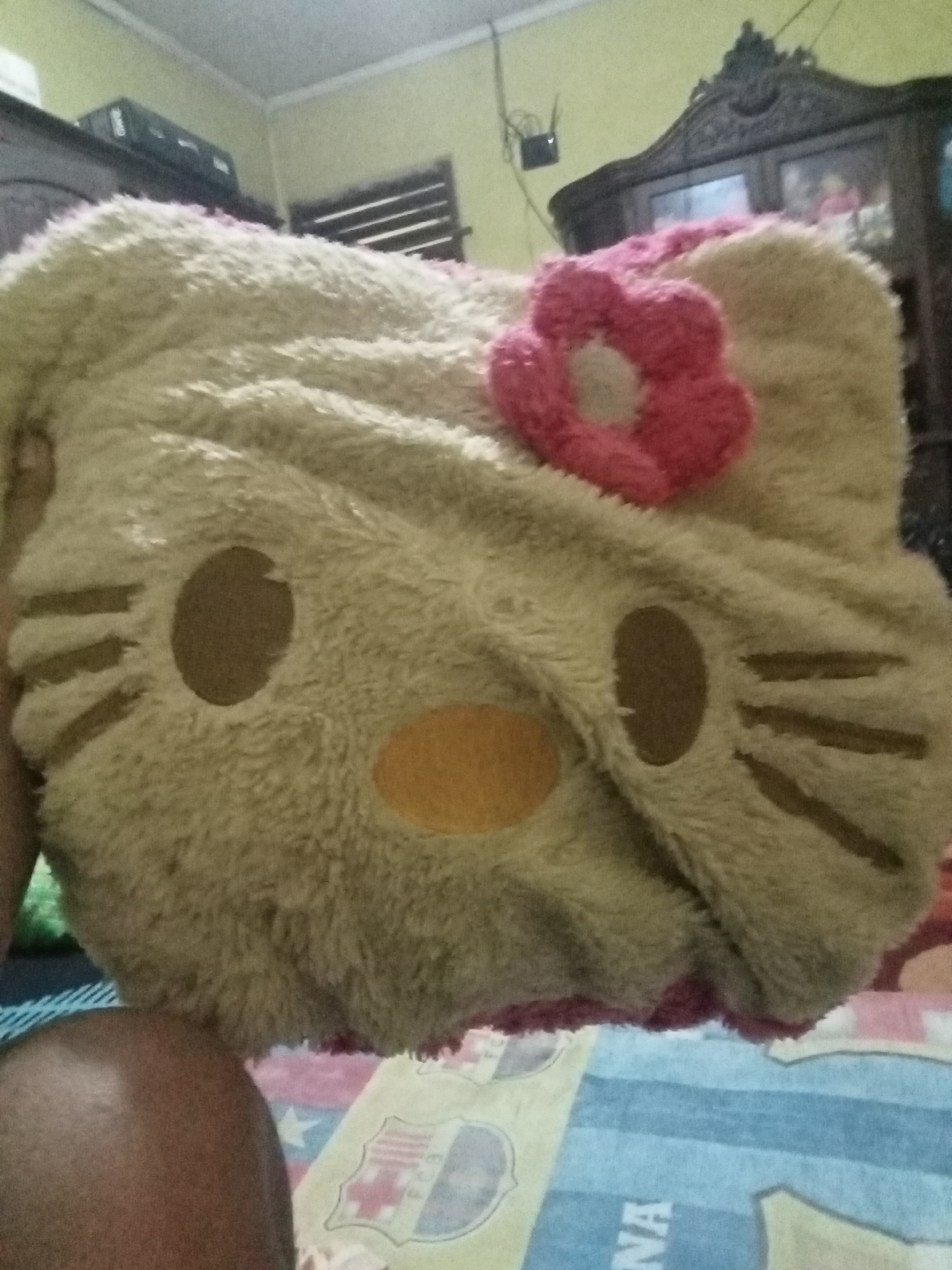 The image depicts a plush, light brown Hello Kitty-shaped pillow with a pink flower accent. The pillow is positioned on a bed with a patterned sheet. In the background, a wooden cabinet and a shelf with electronics are visible. The scene appears to be indoors, possibly in a bedroom. The lighting suggests it might be daytime, though the quality of the image makes it difficult to determine definitively. There is no discernible information linking this image to the city of Grabag, Indonesia.