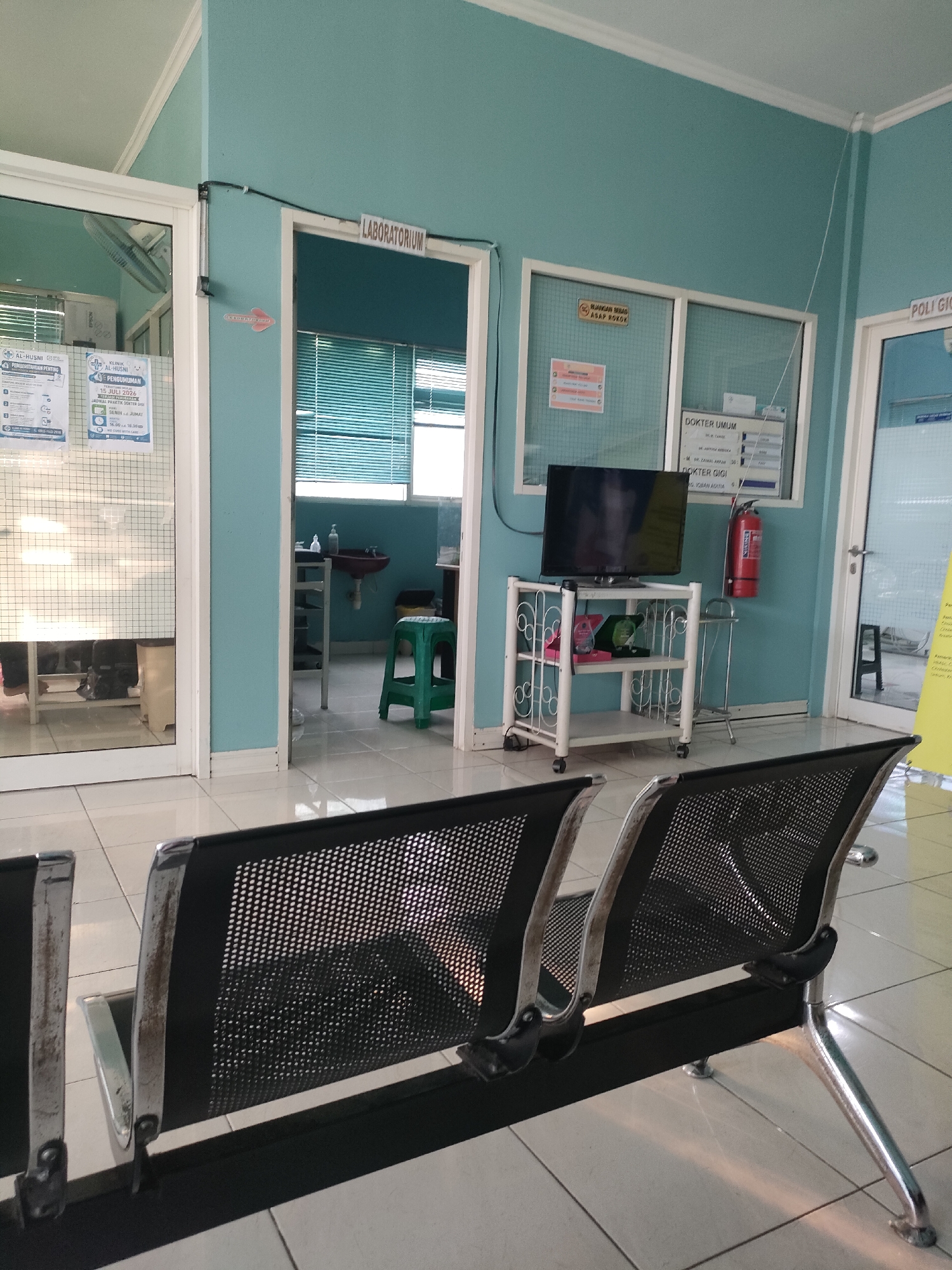 The image depicts a waiting area within a clinic or hospital in Sungailiat, Indonesia. The walls are painted a light teal. In the foreground, two black, metal mesh chairs with chrome legs are visible. To the left, a glass-paneled door reveals a room with a white fan, tables, and posters on the wall. Adjacent to this door, a sign with a red arrow points to "LABORATORIUM". Through the laboratory door, a sink, a green plastic stool, and blinds are visible. Further down the hall, another door is labeled "POLI GIGI". To the right of the laboratory, a white shelf unit holds a television, a fire extinguisher, and other items. A sign above a glass panel reads "DOKTER UMUM" and another beneath it reads "DOKTER GIGI", indicating these are general practitioner and dentist rooms, respectively. A yellow poster is partially visible through the Poli Gigi door. The floor is tiled with a white and beige pattern. Sunlight streams in from the right, casting distinct shadows. There are no people visible in the image.