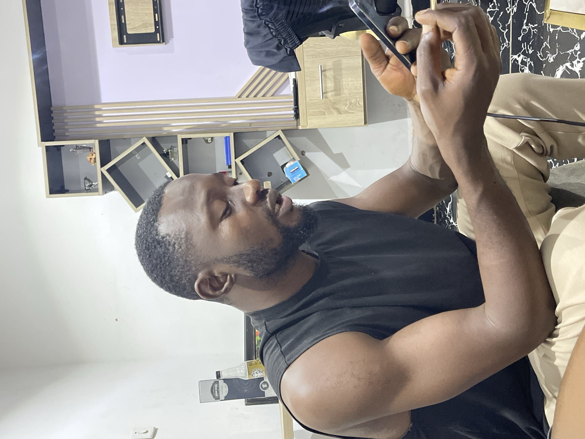 In a modern interior, possibly located in Abuja, Nigeria, a man with dark skin and a beard, wearing a black sleeveless shirt and tan pants, is seated and looking at his mobile phone. He is holding the phone in his right hand, with his left hand positioned to interact with the screen. The background features a wall with a light purple tint, a shelf unit with several geometric compartments, and wooden cabinetry. One compartment on the shelf holds decorative items, while another displays a bottle and a box labeled "John Wick Whiskey." A black cord runs down from the man's lap to the floor. The lighting in the room is ambient, with a slight glow from the phone screen.