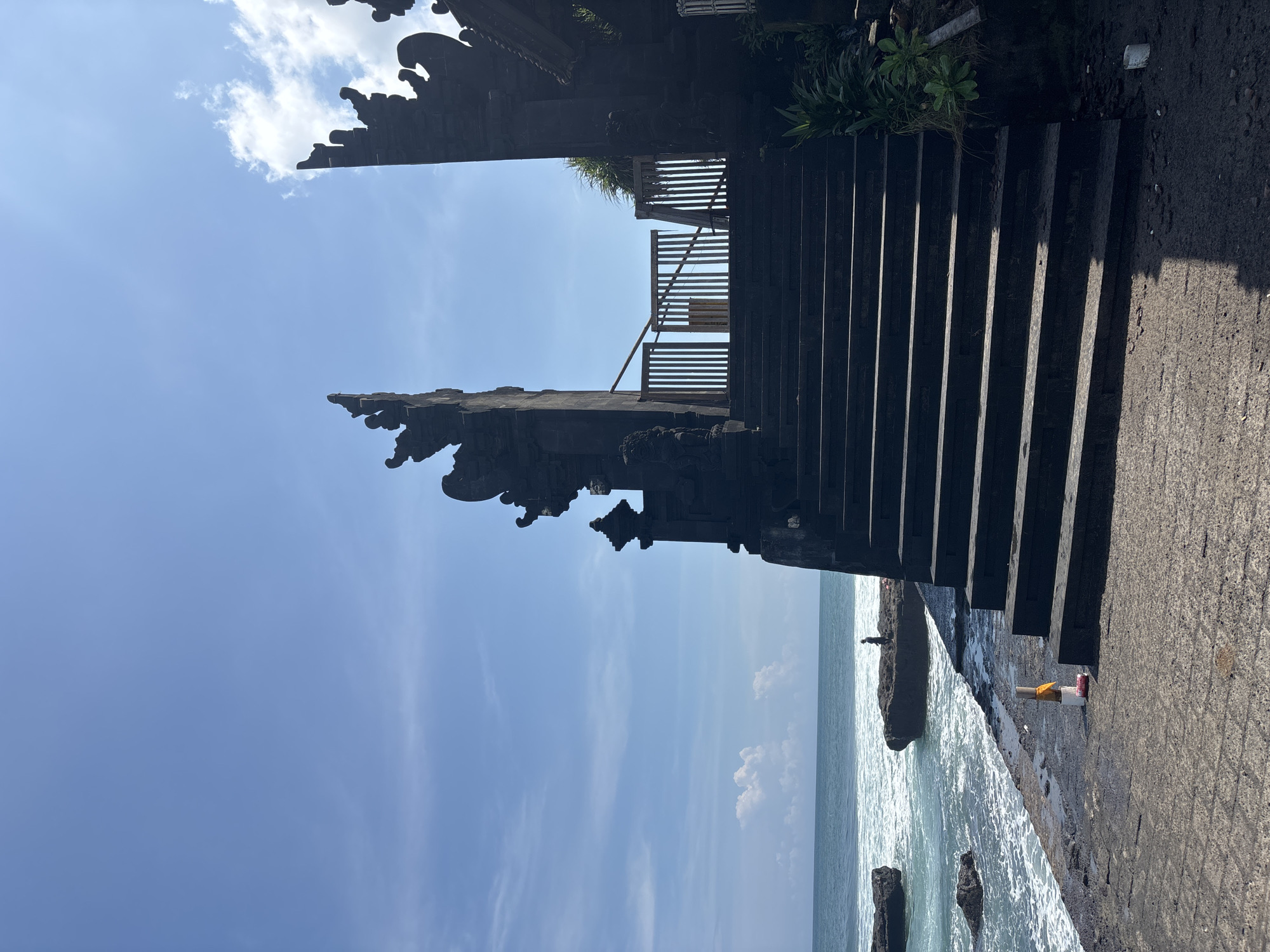 The image depicts a coastal scene in North Kuta, Indonesia. A stone structure with ornate carvings frames the left side of the view, reaching towards a blue sky with scattered clouds. Adjacent to this, a set of dark stone stairs leads upwards, passing a wooden railing. To the right, a concrete walkway is visible, ending at the edge of the ocean. The sea is visible, with waves crashing against rocks. A cluster of discarded trash rests near the edge of the walkway by the steps.