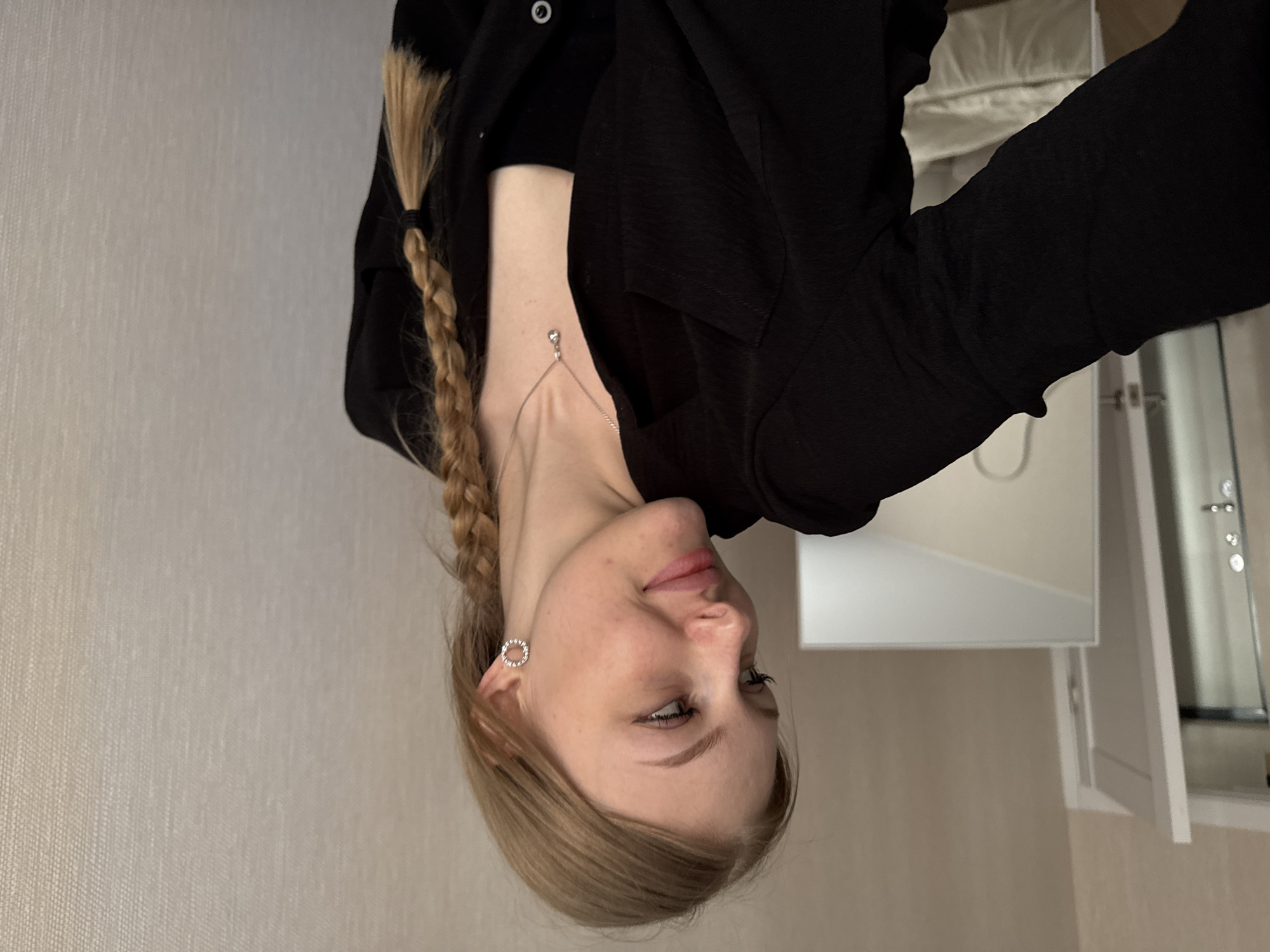 A blonde woman with a braid is posing indoors. She is wearing a black jacket and a silver necklace and earrings. The location appears to be a room with light-colored walls and a white cabinet or vanity. It is not possible to determine the specific location within Perm, Russia from the provided image.