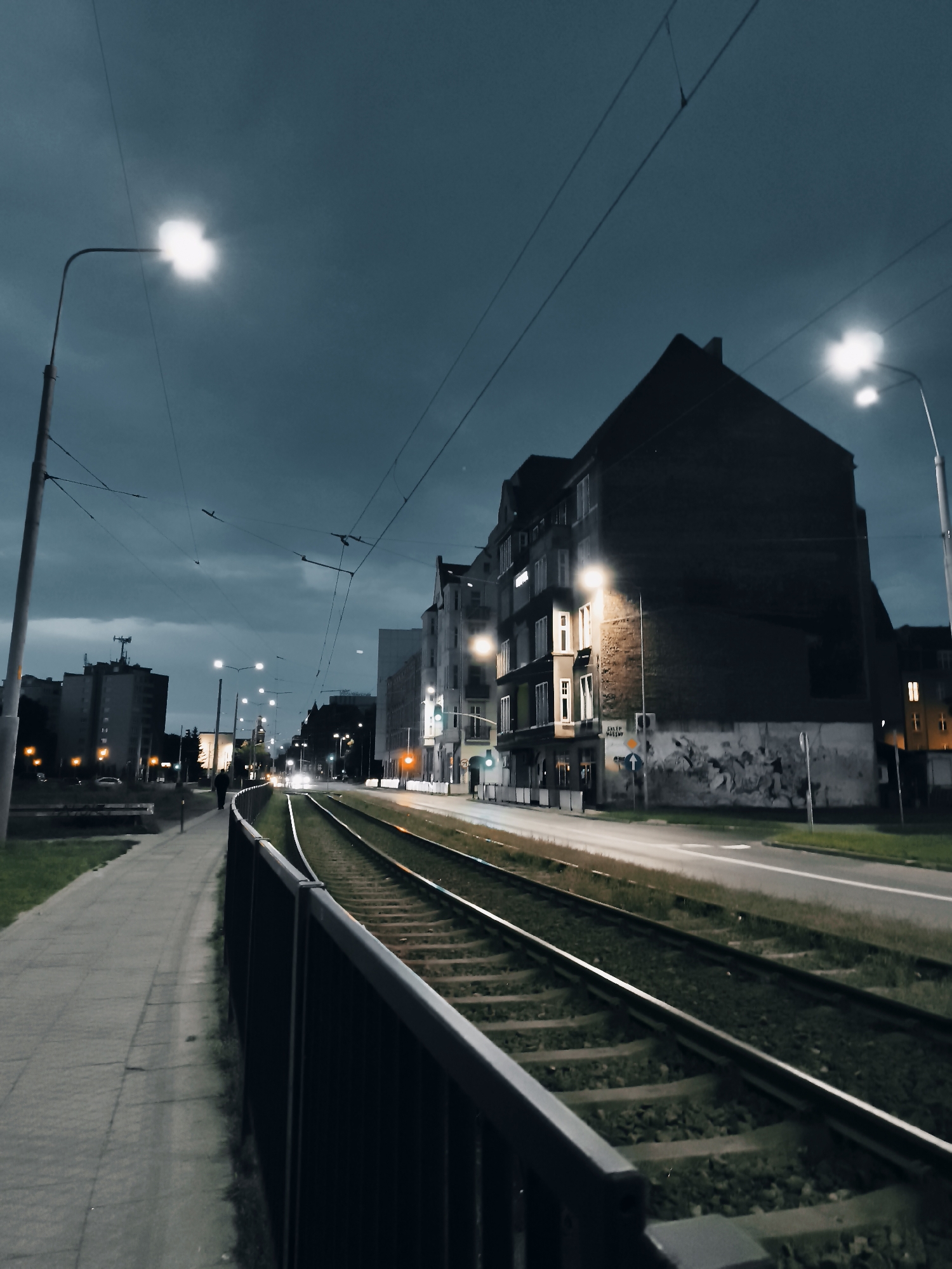 An exterior, low-angle, wide shot captures a tram line and sidewalk in Gdansk, Poland, at dusk or nighttime. The sky is dark and overcast, with a few bright streetlights illuminating the scene. On the left, a sidewalk made of paving stones runs alongside a grassy area. A dark metal fence separates the sidewalk from the tram tracks. The tram tracks themselves are visible in the center of the frame, running parallel to the road. On the right side of the frame, there are several buildings. The most prominent building is a large, dark brick structure with many windows, some of which are lit. A mural is visible on the side of this building. Further down the street, more buildings and streetlights can be seen. A few cars are visible in the distance on the road. A single person is walking on the sidewalk in the distance, away from the camera. The overall mood of the image is quiet and somewhat melancholic, with the artificial lights cutting through the darkness.