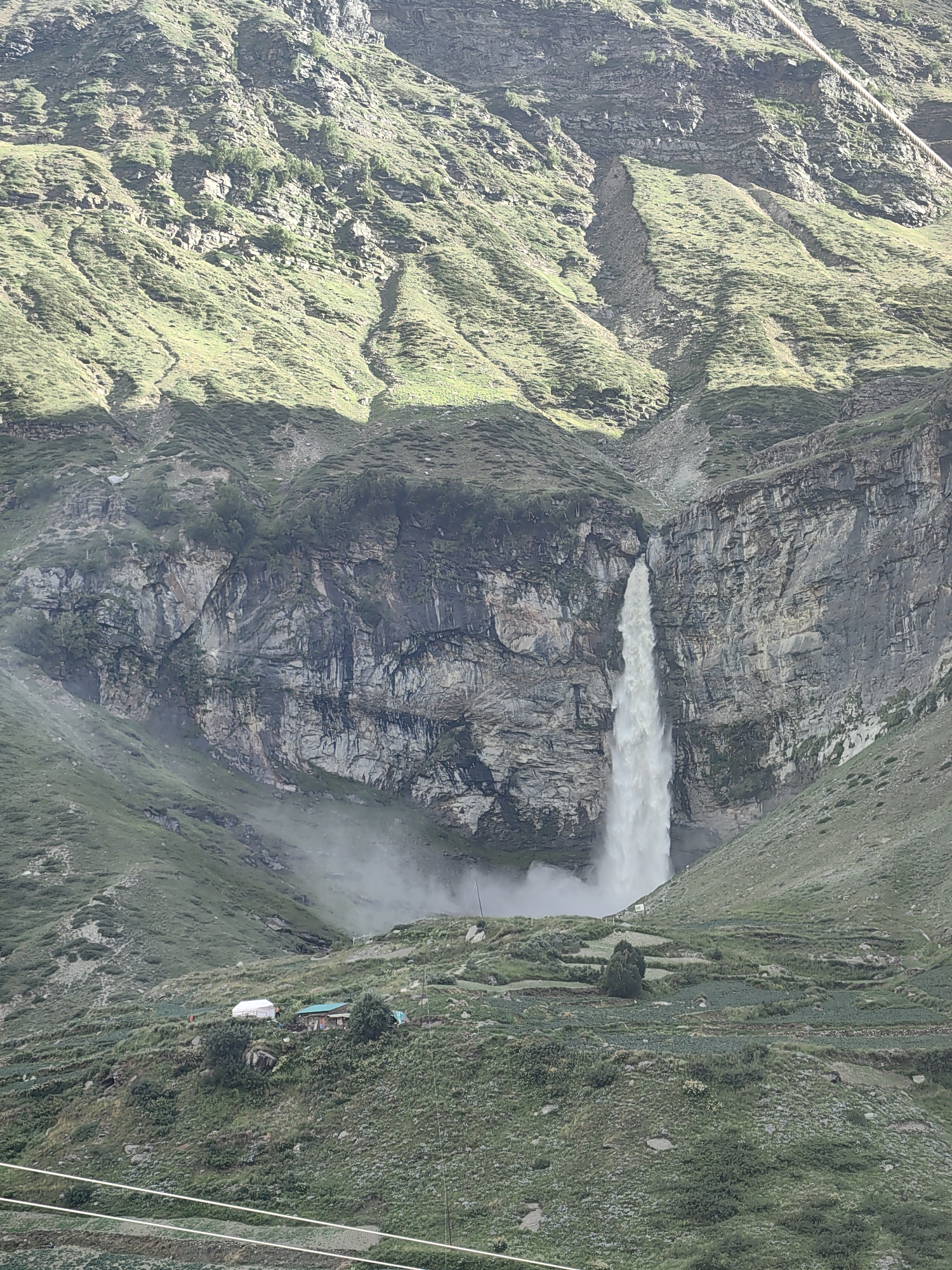 The image depicts a majestic waterfall cascading down a sheer cliff face in the mountainous terrain of Yamling, India. The waterfall is the central focus, with its white water creating a dramatic contrast against the rugged, grey rock of the cliff. The surrounding landscape is characterized by steep, green-covered slopes that appear terraced in places, suggesting agricultural activity. A small cluster of buildings, including a white tent and a house with a green roof, is visible on one of the lower slopes, nestled amongst the vegetation. A fine mist generated by the waterfall hangs in the air near the base of the falls and on the surrounding slopes. The sky above is overcast, casting a diffused light over the scene. The overall impression is one of natural grandeur and remote, rustic habitation.