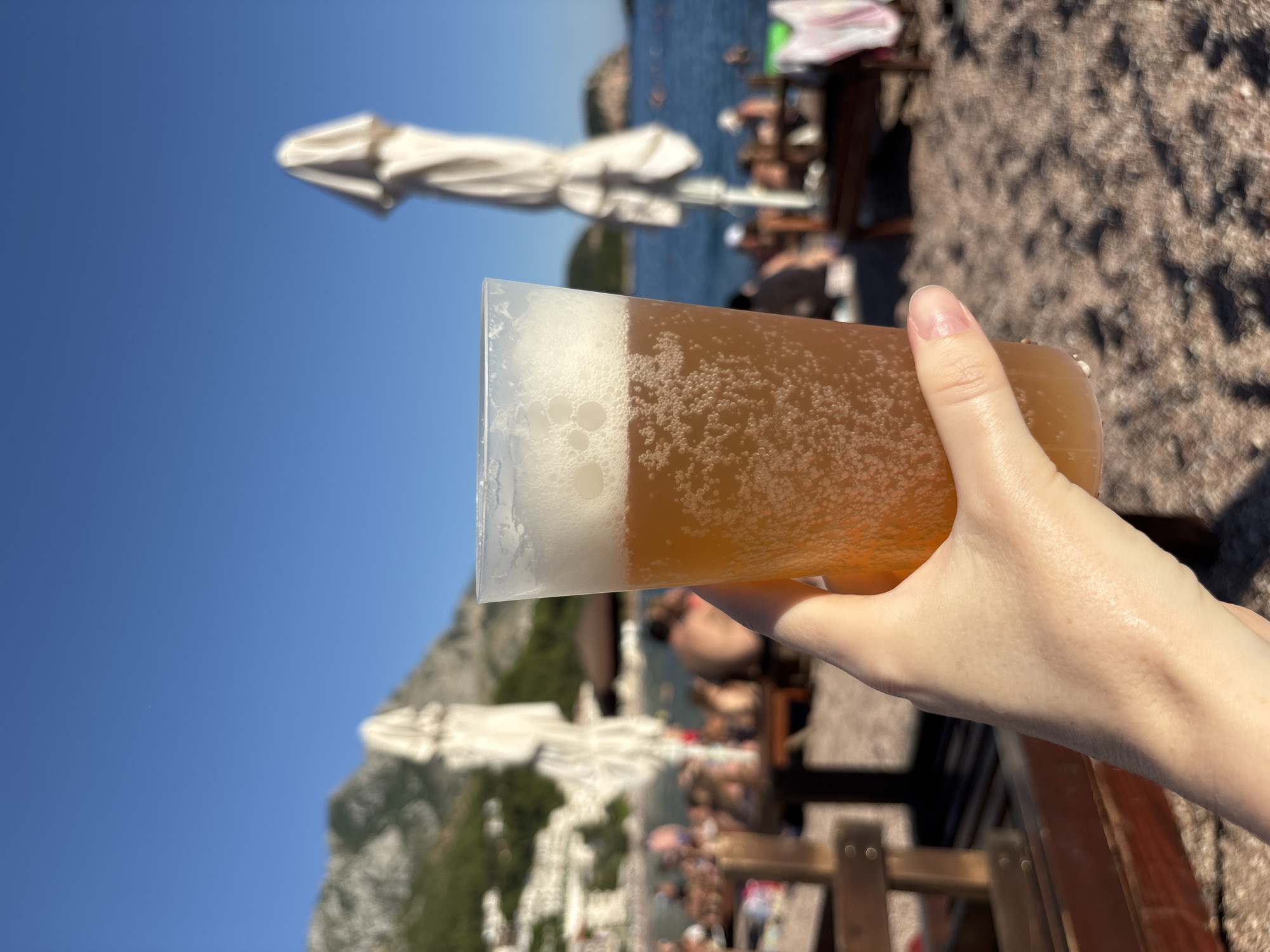 In the foreground, a hand holds a plastic cup filled with amber-colored beer, complete with a frothy head and carbonation bubbles, against a bright blue sky. In the mid-ground, a sandy beach is dotted with sunbathers and beach chairs, with several white beach umbrellas folded and closed. The sea is visible behind the beach, with faint shapes of people swimming. In the background, lush green hills rise, with buildings nestled into the slopes, suggesting a coastal town or resort. The overall setting is a sunny day at the beach in Sveti Stefan, Montenegro, a popular tourist destination known for its scenic coastline and historical island.