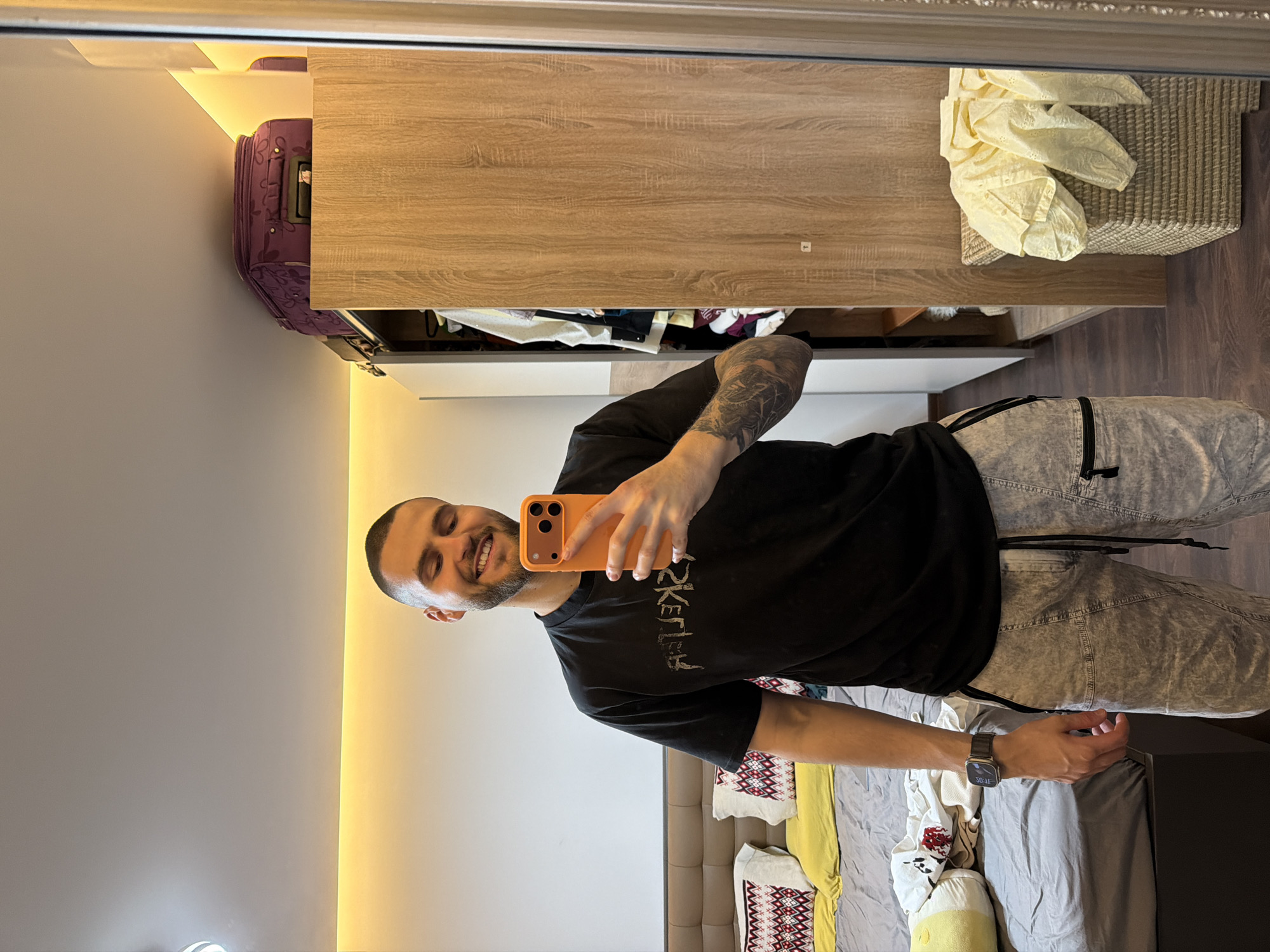 The image shows a man taking a selfie in front of a mirror. He has short hair and a beard. He is wearing a black t-shirt, light gray pants, and a wristwatch. He is holding an orange smartphone. Behind him is a bedroom setting with a bed, pillows, a wardrobe, and a suitcase on top of the wardrobe. It is not possible to determine the exact location in Sofia, Bulgaria from the image.