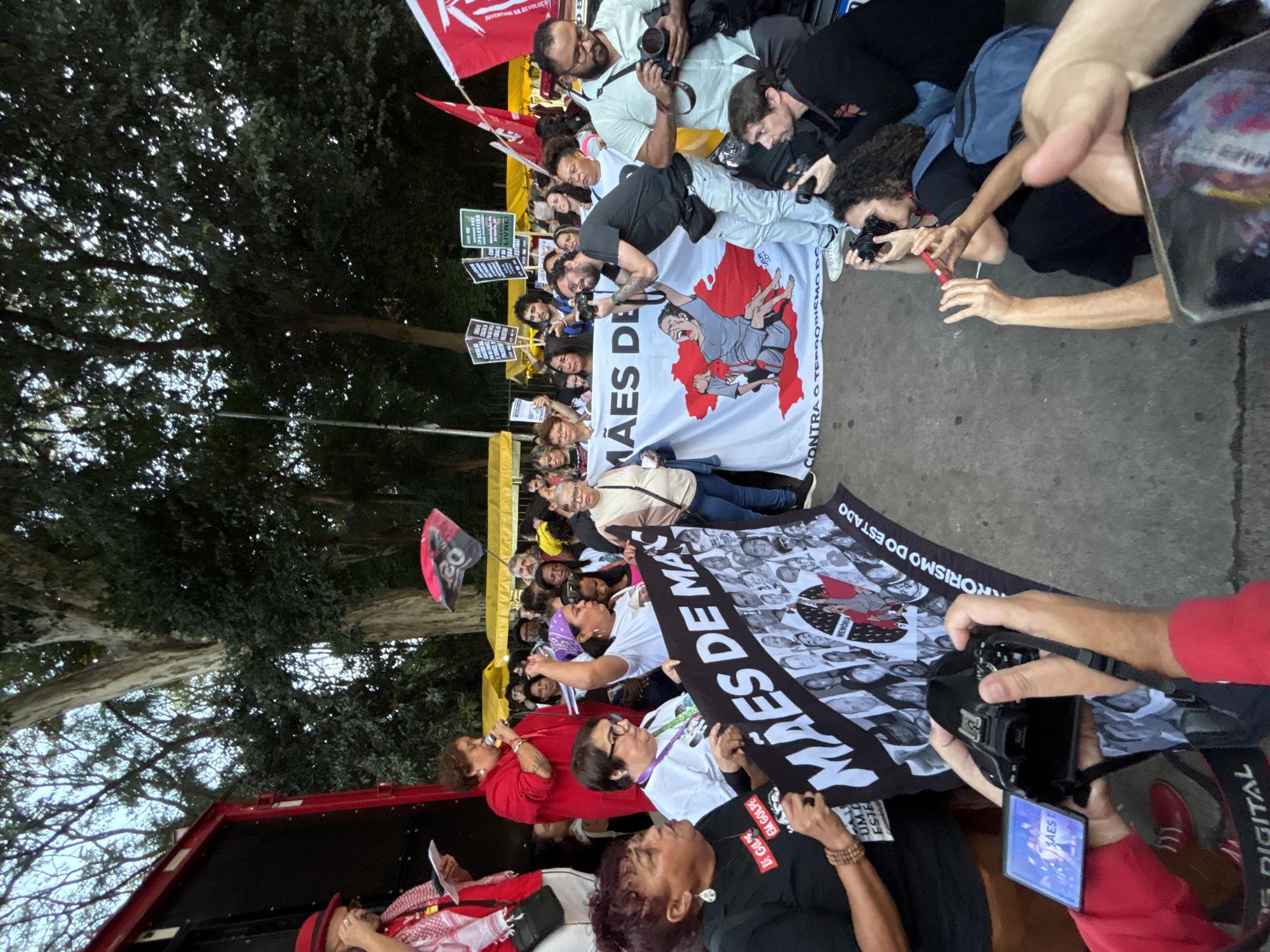 A street demonstration takes place with participants holding banners and signs. A large banner reads "Mães de Maio contra o terrorismo do Estado" (Mothers of May against state terrorism) with a cartoon depiction of someone being tortured and with photos of people. On the right, people are taking pictures of the protest with cameras and phones. Trees can be seen in the background, along with yellow and red flags. The location may be the city of São Paulo, Brazil.
