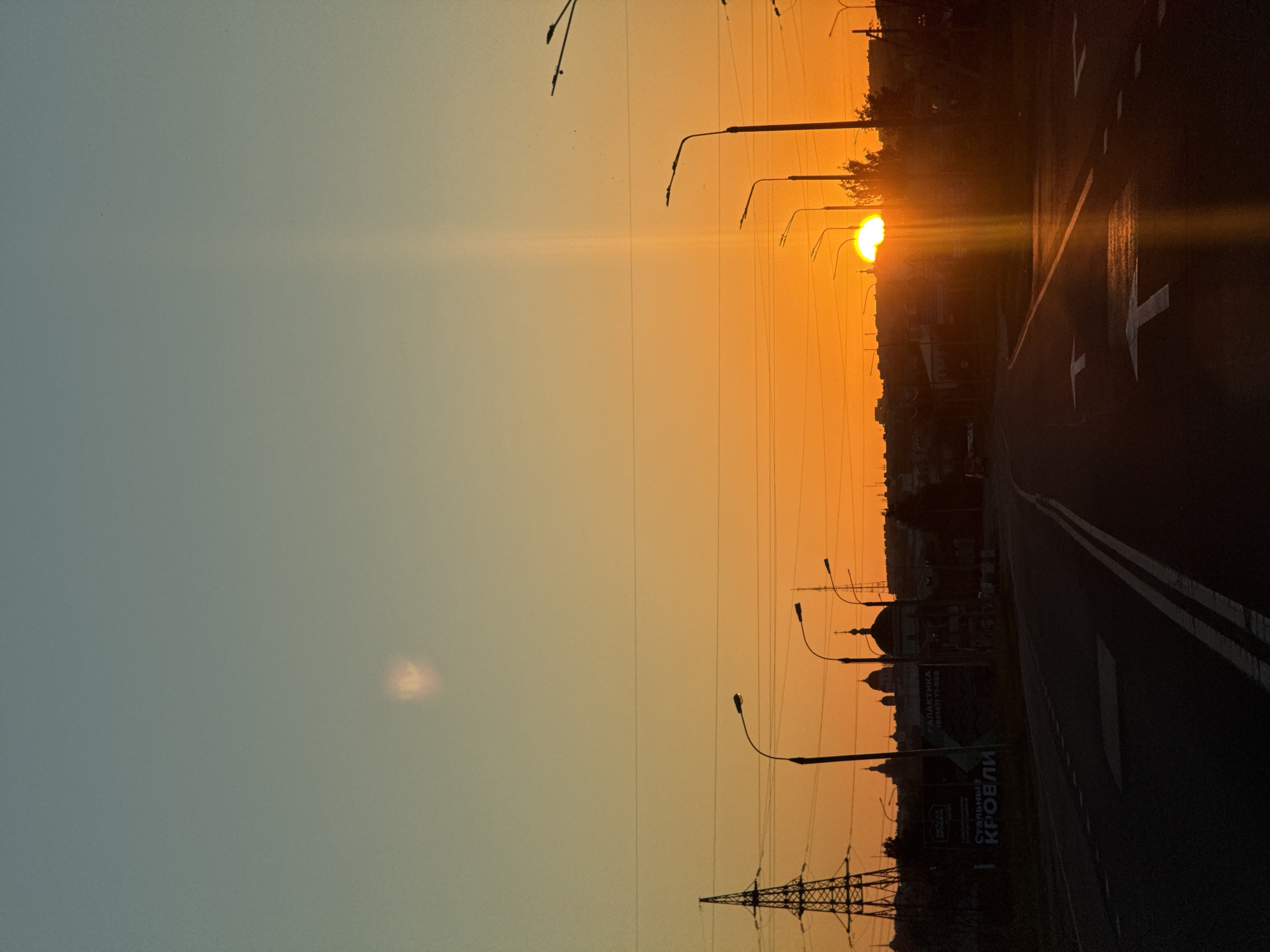The image shows a sunset over a highway in Morózovka, Russia. The sun is setting behind a silhouette of buildings and trees, casting a warm orange glow across the sky. The sky transitions from a soft blue at the top to a vibrant orange near the horizon. In the foreground, the highway is dark with visible lane markings. Several streetlights line the right side of the road, their silhouettes stark against the bright sky. A large power pylon is visible on the left side of the image. The overall scene depicts a serene and picturesque view of a sunset in a semi-urban environment.