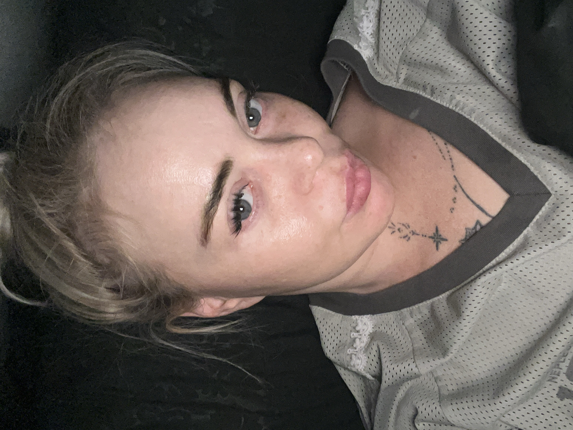 A close-up, overhead shot shows a young woman with blonde hair pulled into a messy bun against a dark background. She is wearing a light gray athletic jersey with dark gray trim around the V-neck. The woman has light blue eyes, prominent eyelashes, and fair skin with some redness and freckles. Her lips are slightly parted, and she is looking downwards. A tattoo is visible on her upper chest, appearing to be a constellation or a star. The lighting is dim, highlighting the sheen on her face. The location is unspecified.