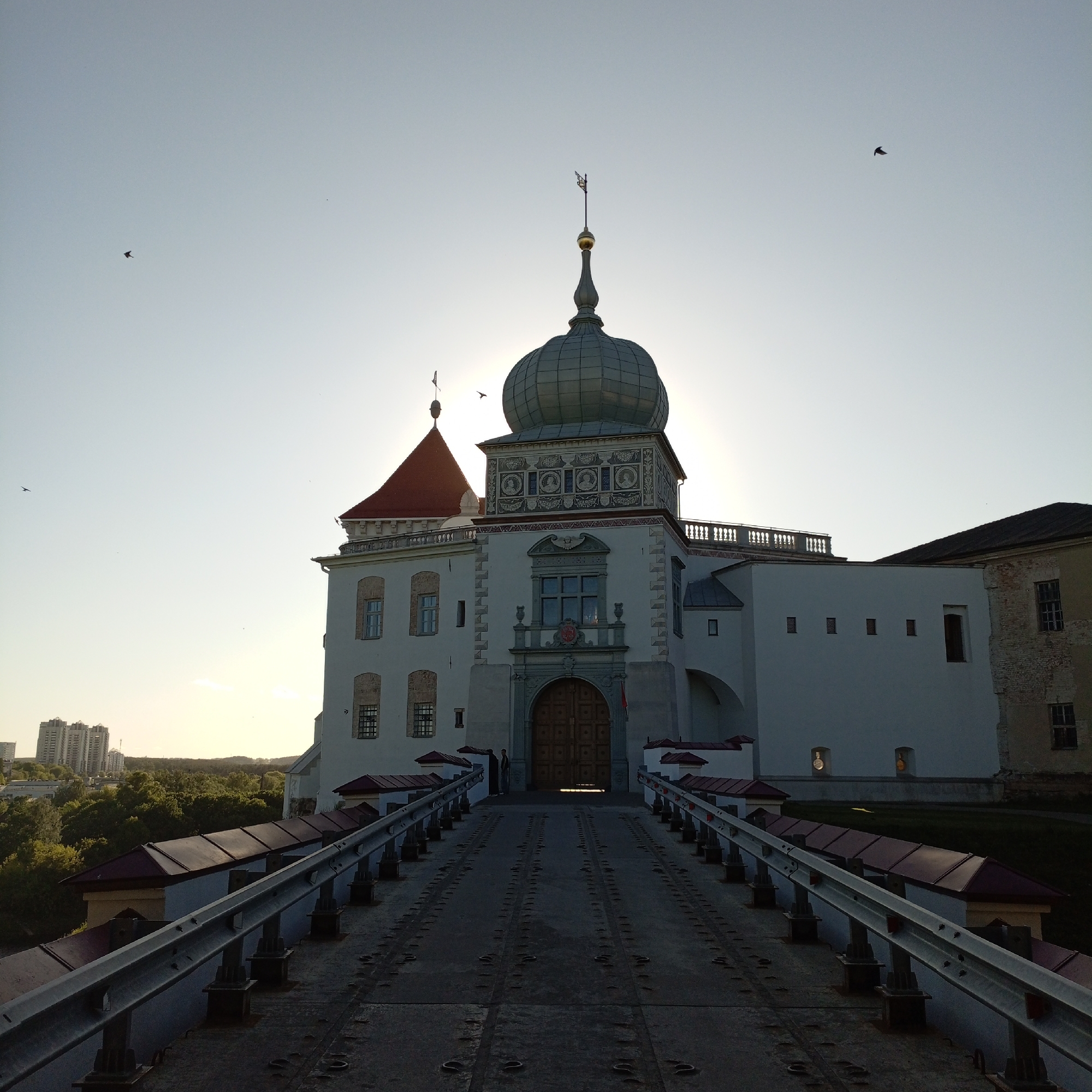 The image shows a castle with a bridge leading up to its main entrance. The castle has a distinctive green dome and a red-roofed section. The sun is behind the castle, creating a halo effect. A few birds are visible in the sky. In the distance, some buildings and trees can be seen. The bridge is made of metal and has railings on both sides. A figure appears to be standing in the castle's doorway. The location appears to be a cityscape, with the castle as a prominent historical feature.