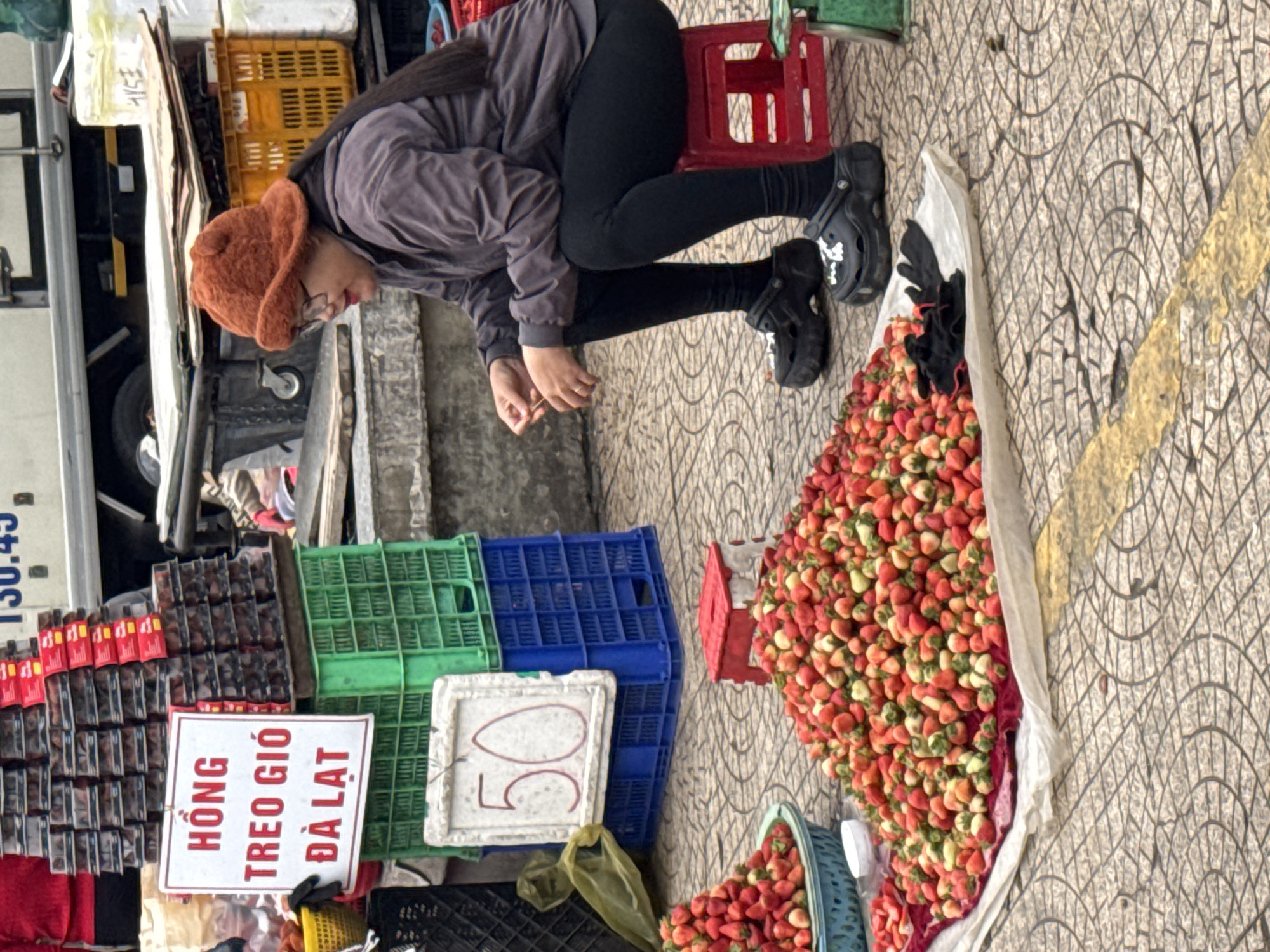 In Da Lat, Vietnam, a woman wearing a brown fuzzy hat and glasses is sitting on a small red stool at an outdoor market. She is wearing a dark gray jacket and black leggings, with black chunky clogs on her feet. In front of her, a large pile of fresh strawberries is spread out on a white cloth on the ground. To her left, there are several crates filled with strawberries, and a sign in Vietnamese that reads "Hồng treo gió Đà Lạt" (wind-dried persimmons from Da Lat), although the fruit appears to be strawberries. Another sign with the number "50" is visible behind the crates. In the background, a truck and more stacked crates suggest a busy market environment. The ground is paved with textured tiles.