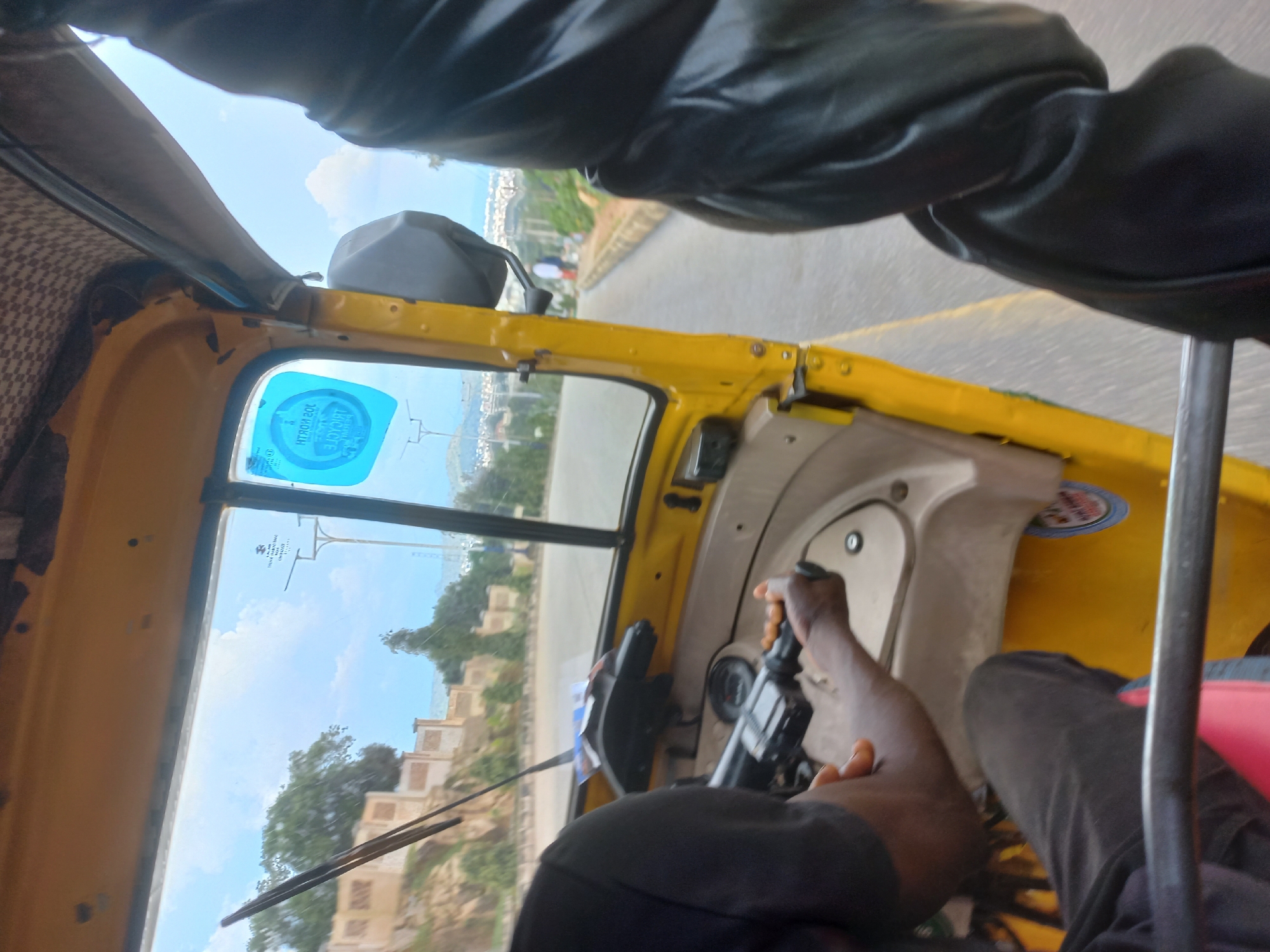 The image is taken from the interior of a yellow auto-rickshaw, looking out through the windshield. The driver, a person of color wearing a black t-shirt and dark pants, is visible from the shoulders down, with their hands on the steering mechanism. The auto-rickshaw's interior is visible, including the yellow dashboard and a black seat. Outside the auto-rickshaw, a road is visible, with a sidewalk and a gentle slope on the left side of the frame. Trees and buildings are visible in the distance, suggesting an urban or semi-urban environment. The sky is bright and partly cloudy. There is a blue sticker on the windshield, which appears to have some text and graphics on it. The overall impression is a candid shot taken while traveling in an auto-rickshaw.