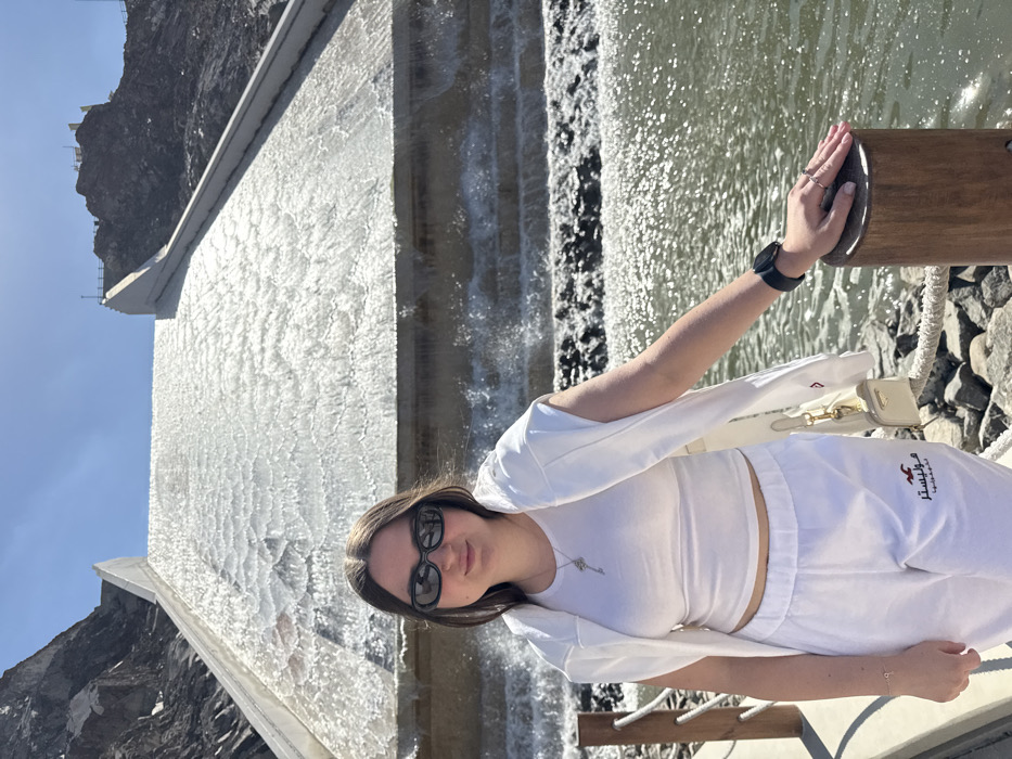 The image displays a young adult female standing outdoors at the Hatta Dam spillway in Hatta, United Arab Emirates, under a clear blue sky. The female, positioned slightly off-center to the left, has dark hair, fair skin, and wears black oval sunglasses. She is dressed in a white ensemble consisting of a white crop top, a white sleeveless open vest draped over her shoulders, and white sweatpants featuring a "Hollister" logo with Arabic script. Accessories include a silver key-shaped pendant necklace, a thin silver bracelet on her left wrist, a black digital watch on her left wrist, and a silver ring on her right ring finger. Her right hand rests on the flat top of a light brown wooden bollard, which is part of a rope railing system in the immediate foreground.
Behind the subject, a wide concrete spillway extends diagonally upward and to the left, covered entirely by a continuous sheet of flowing water that generates ripples and reflections. The water cascades over several tiered steps at the base of the spillway before entering a sun-dappled body of water visible in the lower right and mid-ground. Steep, rocky, barren mountain slopes frame the spillway on both sides and extend into the background. A bright white concrete structure with a railing is visible atop the dam on the upper right side. The scene is brightly illuminated by direct sunlight.