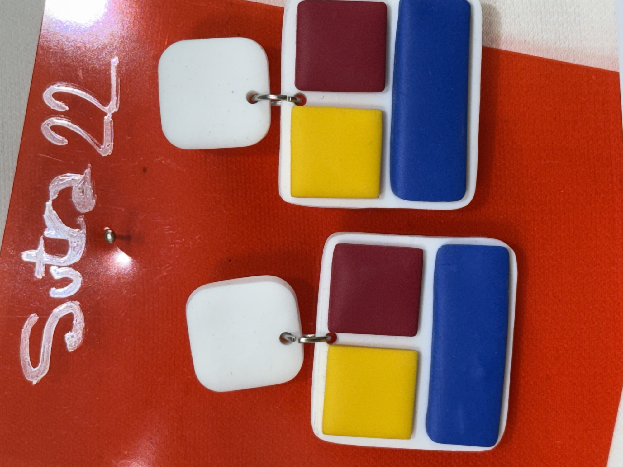 The image shows two handmade earrings laid on top of what appears to be a red surface, possibly a piece of fabric or paper, with white writing on it saying "Suča 22". The earrings are comprised of a white square, connected via a metal loop to a rectangular piece containing a red square, a yellow square, and a blue rectangle. The surface they are lying on is a gradient of red and white. The scene is located in Novi Sad, Serbia because Suča 22 is a street address there.