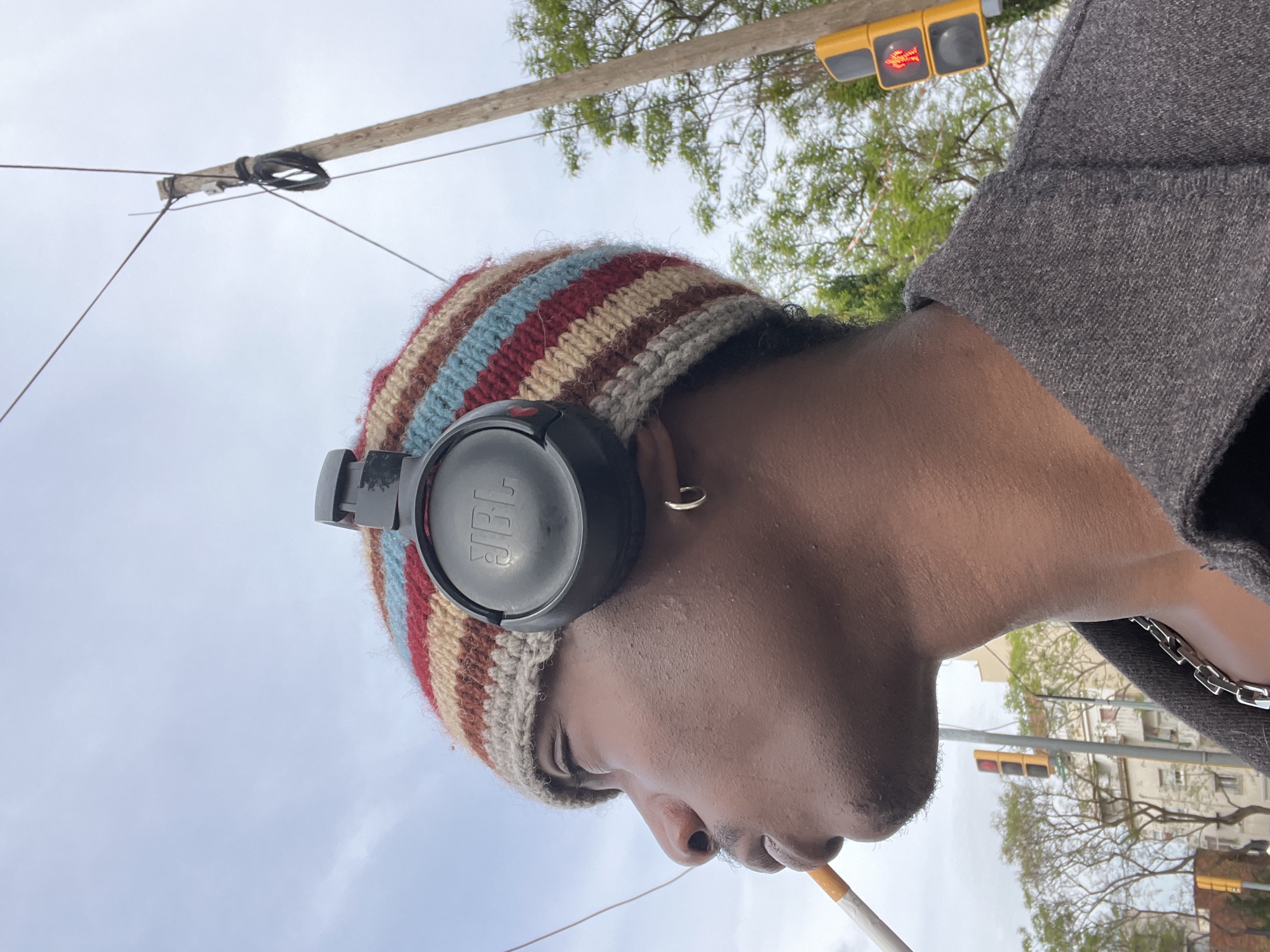 A close-up, low-angle portrait shows a person with dark skin in profile, looking slightly downwards, against an overcast sky. The image is rotated 90 degrees clockwise from a typical upright orientation. The person wears a multi-colored striped knit beanie with black JBL over-ear headphones resting over it. A small golden hoop earring adorns their right ear. A lit cigarette is held at their mouth. They are dressed in a dark gray jacket or coat, beneath which a silver chain necklace is visible.
In the background, urban elements are present: a wooden utility pole with wires spans the upper frame, and a traffic light with both red "Don't Walk" signals illuminated is visible to the right. Green-leafed trees are seen behind the pole and traffic light, and faint light-colored buildings with balconies suggest an urban street setting. The scene takes place outdoors in Barcelona, Spain, under an overcast sky during daytime. The only visible text is the 'JBL' logo on the headphones.